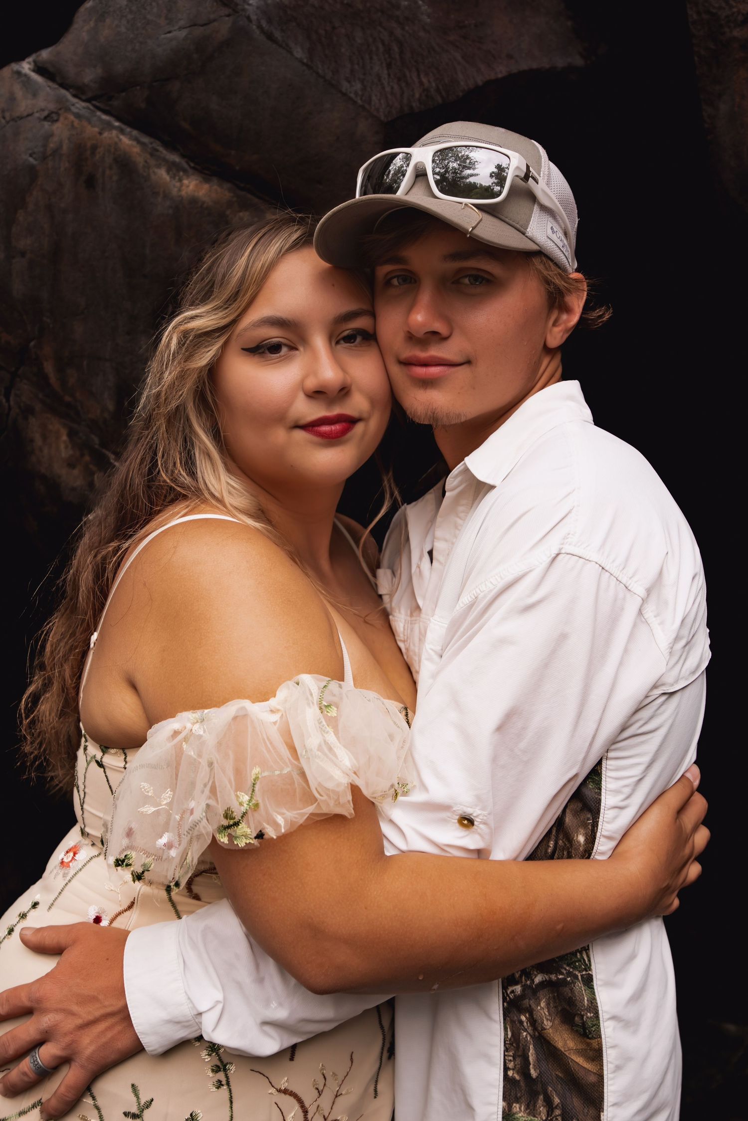 A romantic couple embraces outdoors, with the woman wearing a white off-shoulder dress and the man in casual attire.