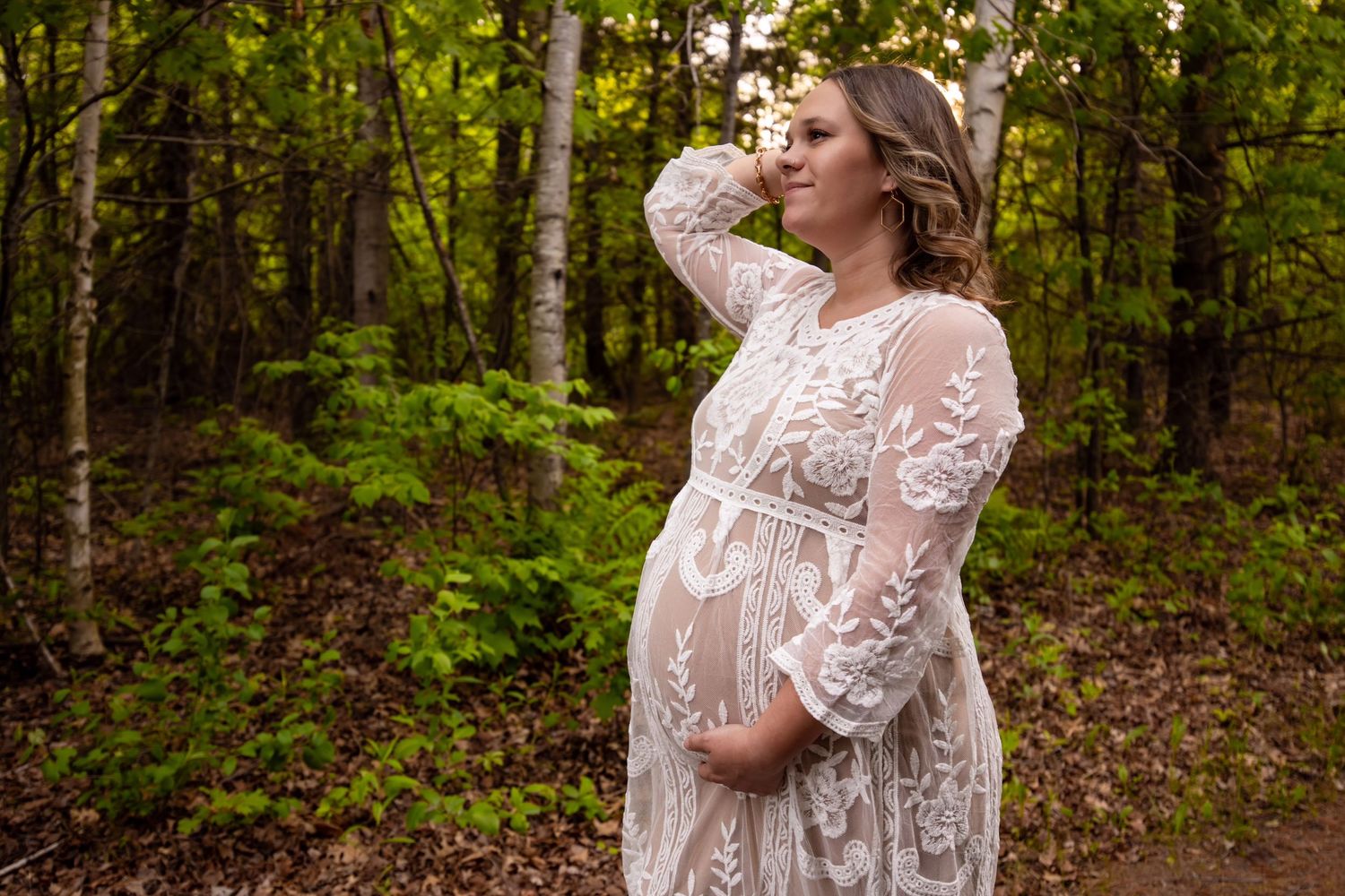 Ethereal maternity portrait in a lace white dress standing in a sunlit forest setting.
