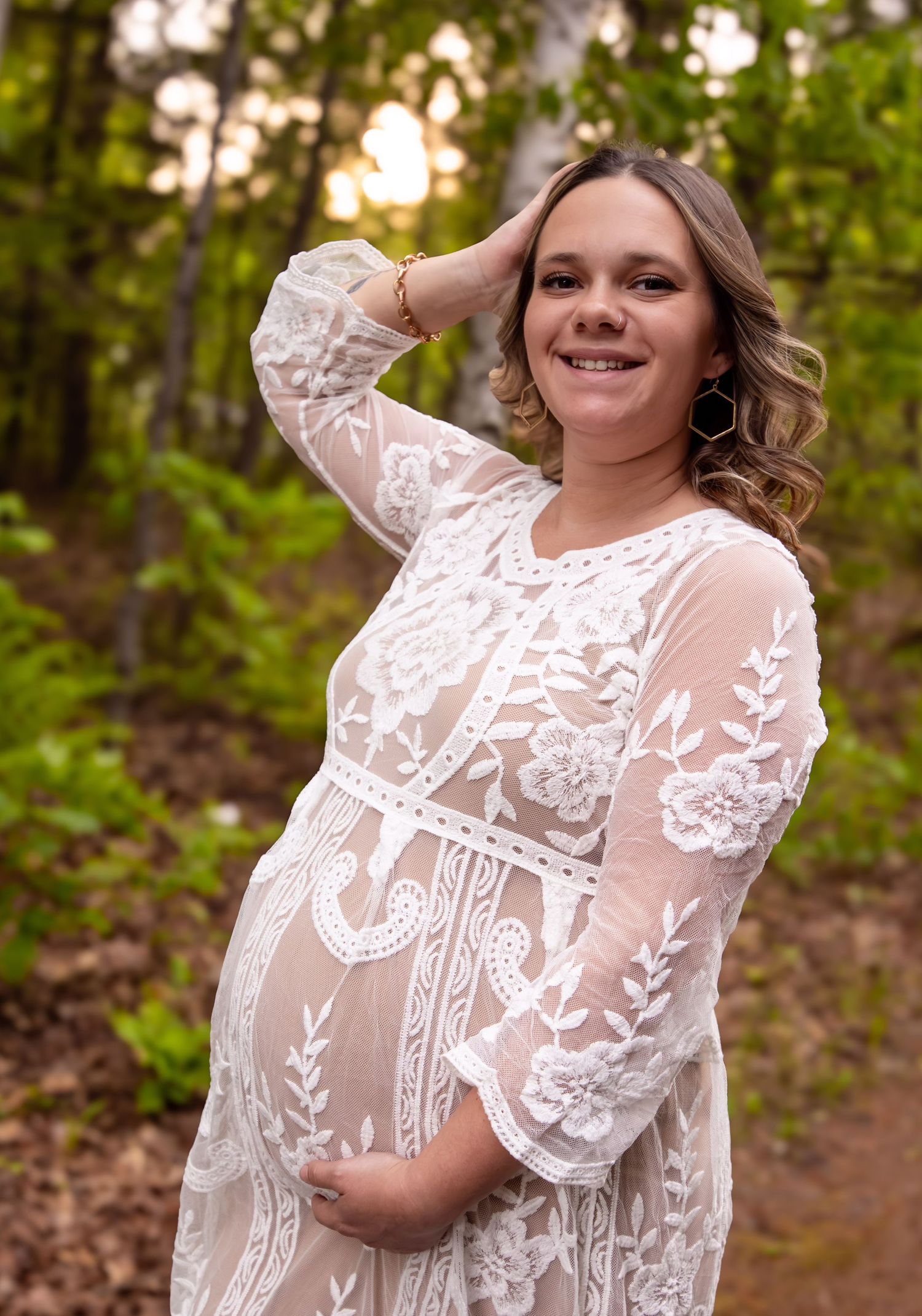 Expectant mother in flowing white lace dress poses joyfully in a wooded setting during golden hour.