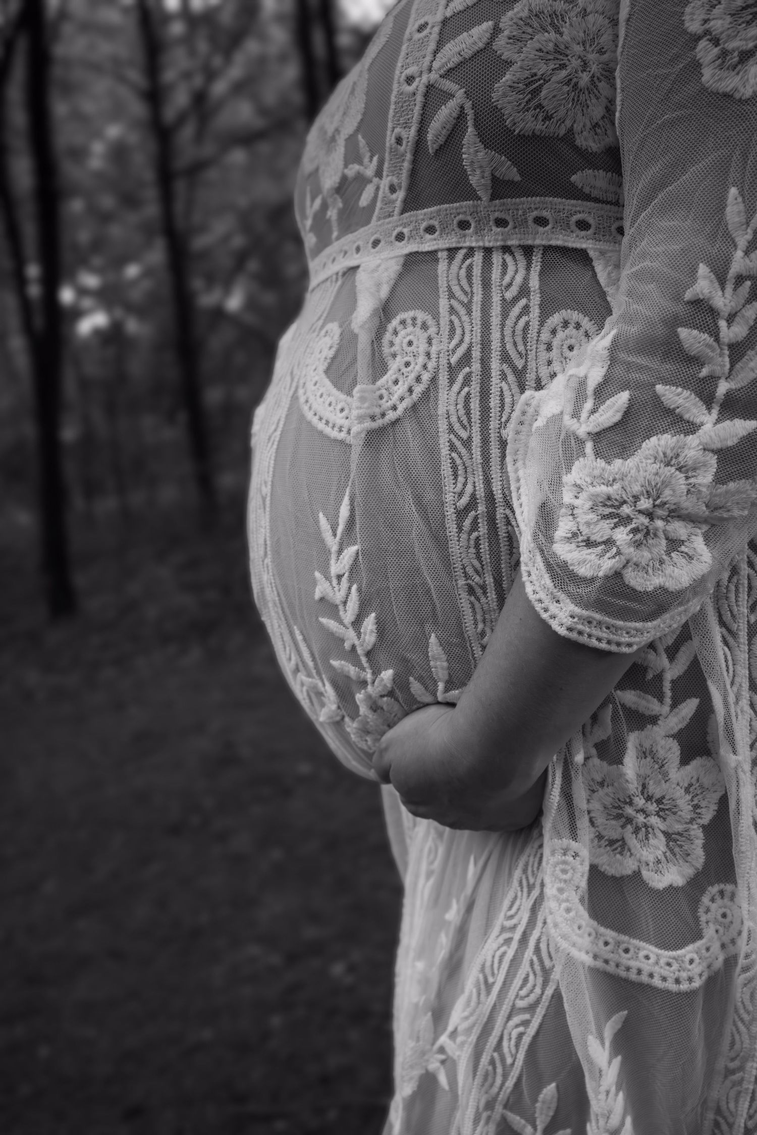 Close-up of an ornate lace maternity dress with floral patterns against a dark forest background.