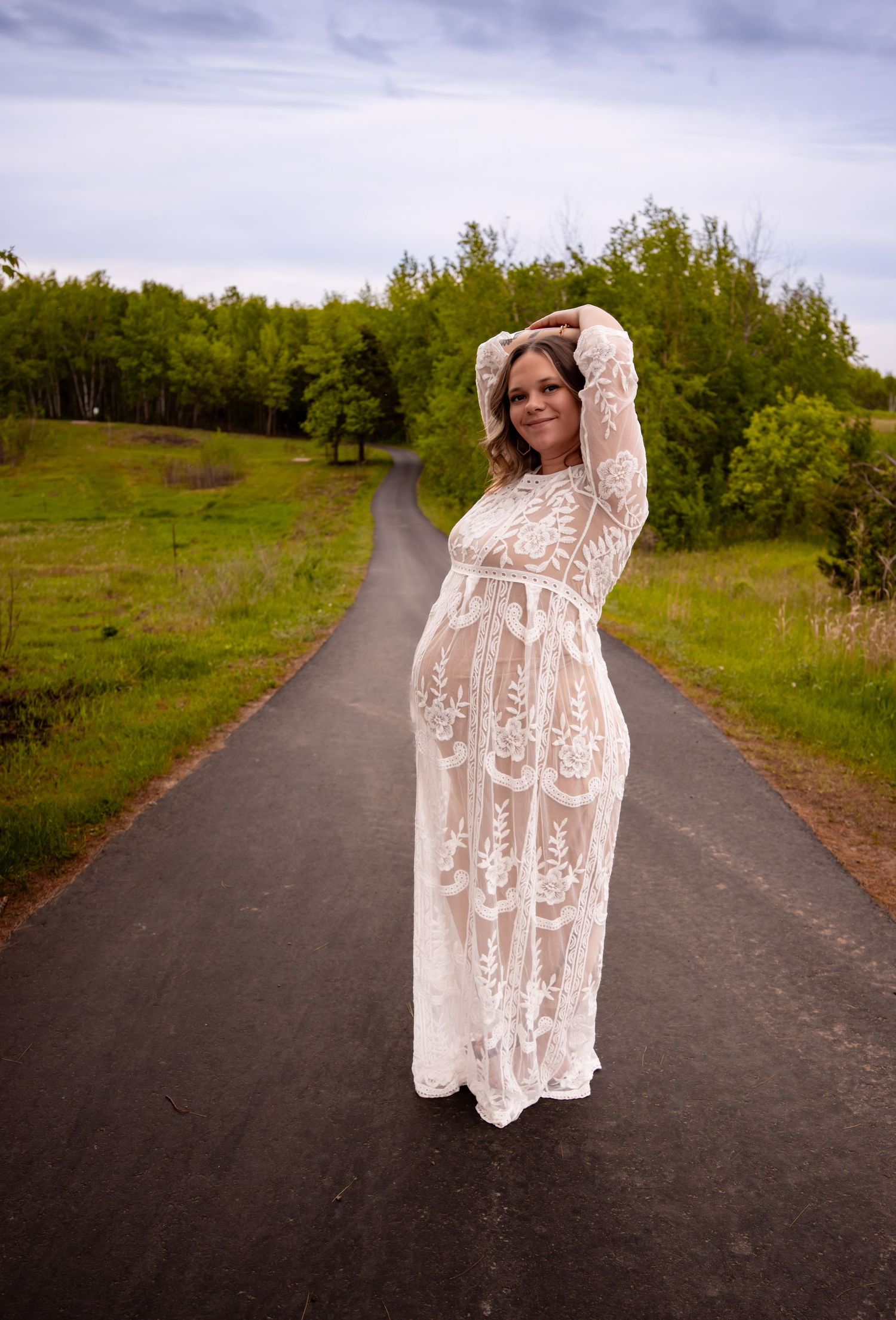 A figure in a flowing white lace dress poses dramatically on a winding paved path surrounded by green foliage.