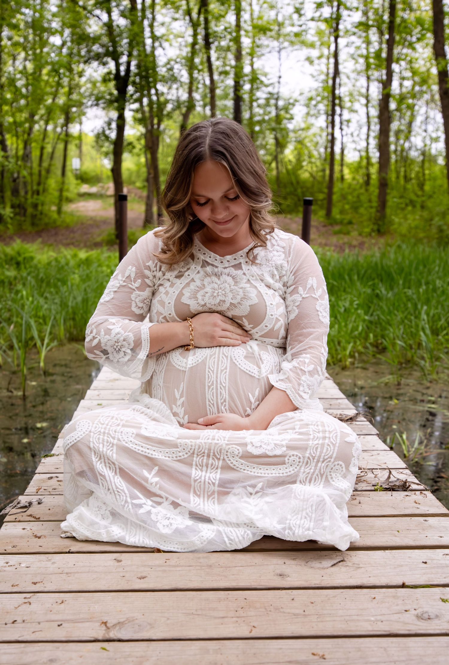 A pregnant woman in a white lace dress sits on a wooden dock surrounded by green trees.