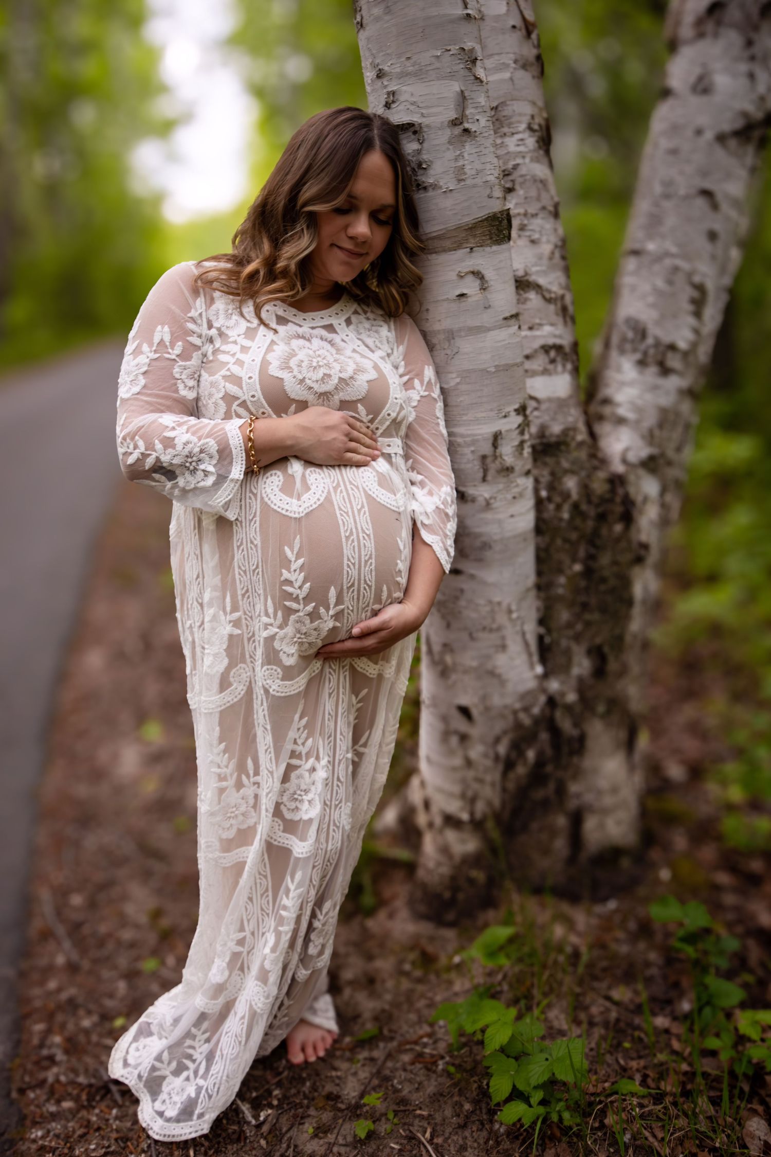 Maternity portraits featuring a delicate white lace dress against birch trees in a wooded setting.