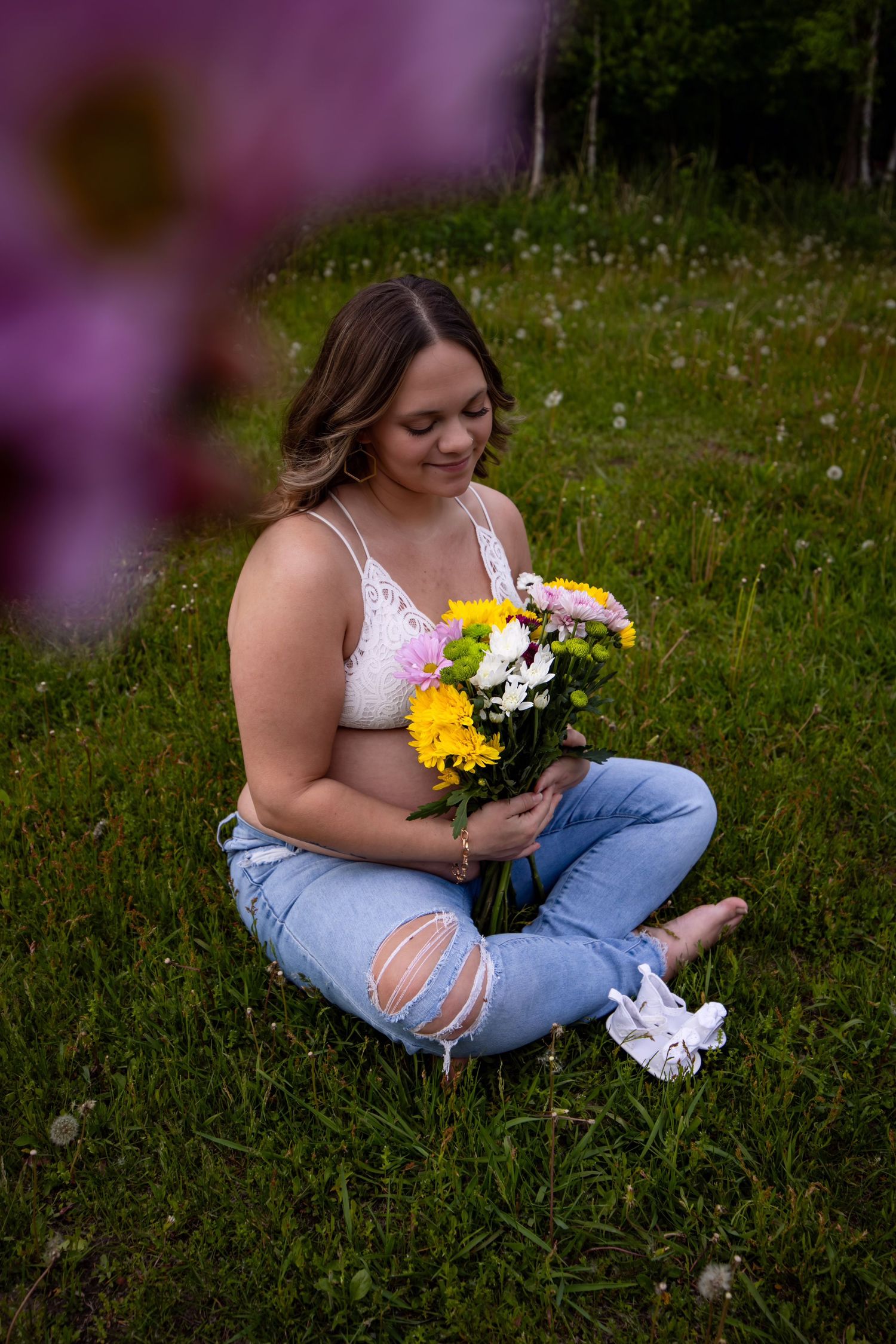 Person in white top and jeans sits in grassy field holding yellow and white floral bouquet.