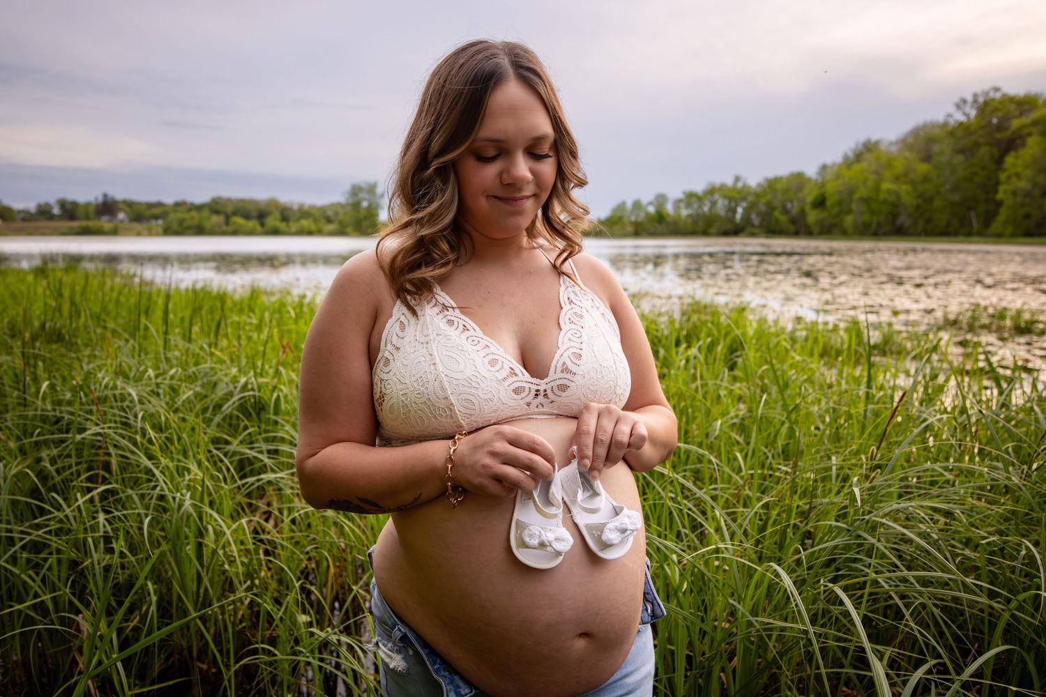 Expectant mother in white dress holds tiny baby shoes while standing in a meadow near a lake at sunset.