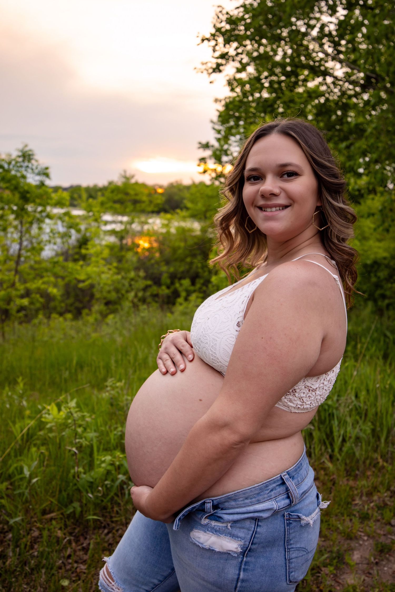 A pregnant woman in white top and denim poses outdoors in a grassy field during golden hour.
