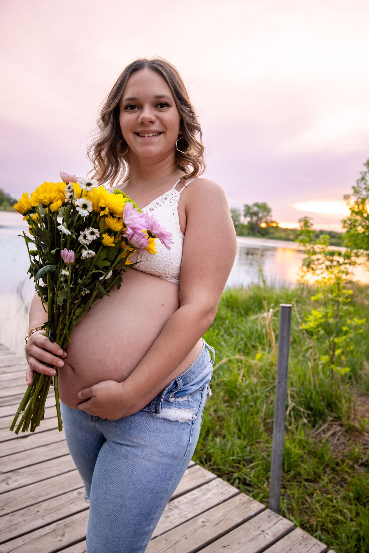 Maternity photo on wooden boardwalk at sunset with person holding bouquet of yellow and pink flowers.