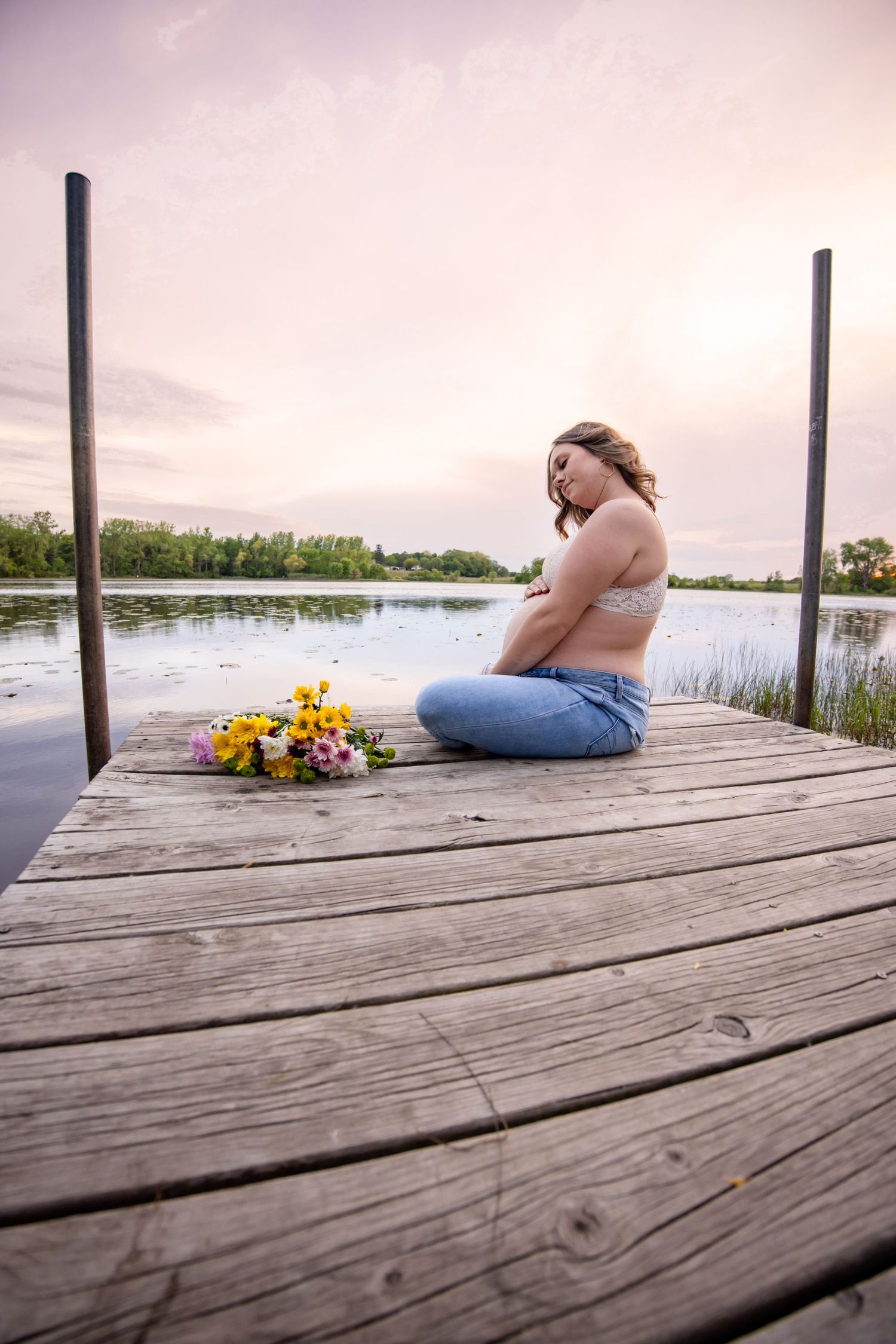 A peaceful maternity moment on a dock with yellow flowers at sunset by a serene lake.