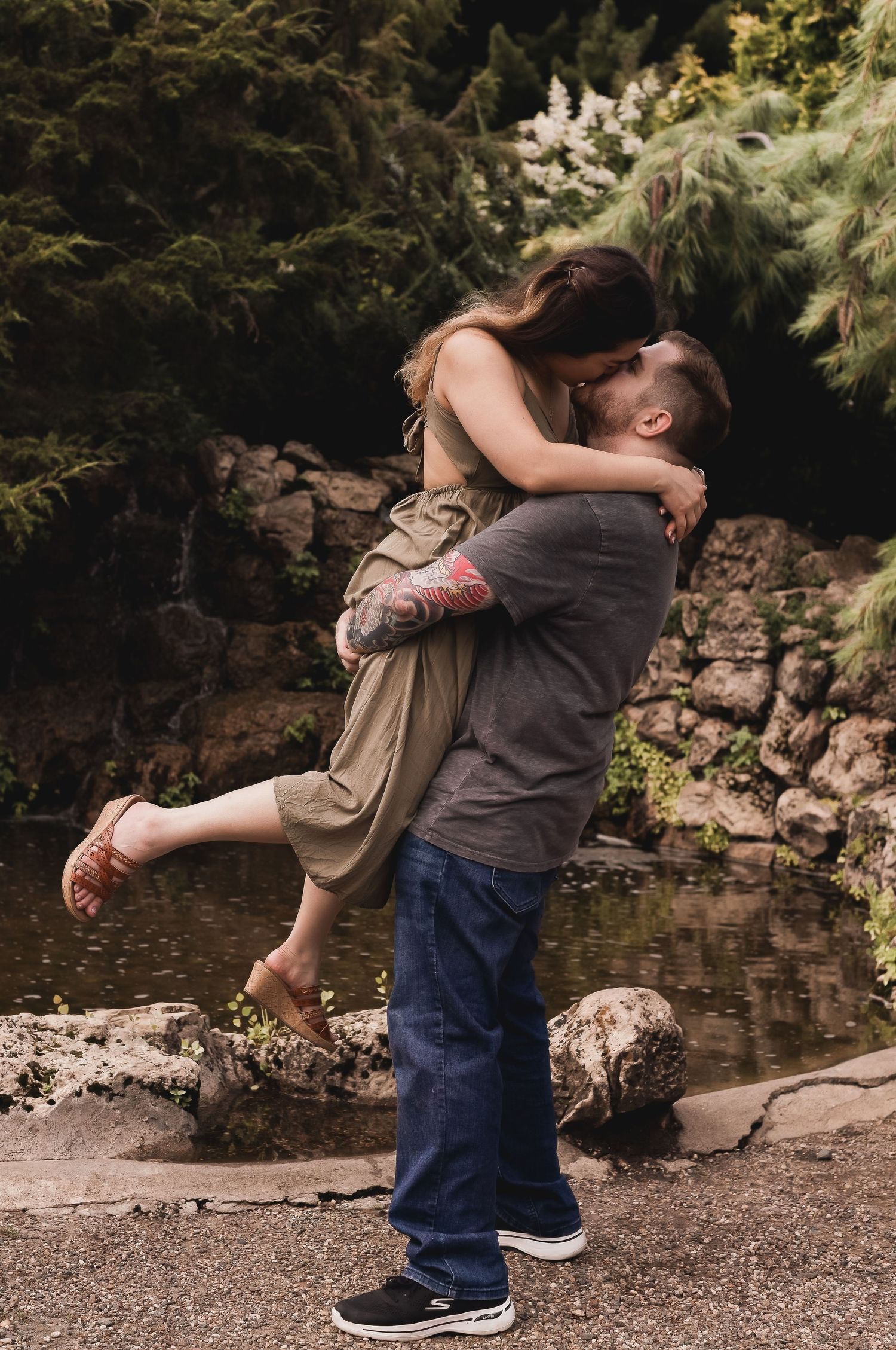 A romantic couple sharing an intimate embrace by a stone wall and pond in a lush garden setting.