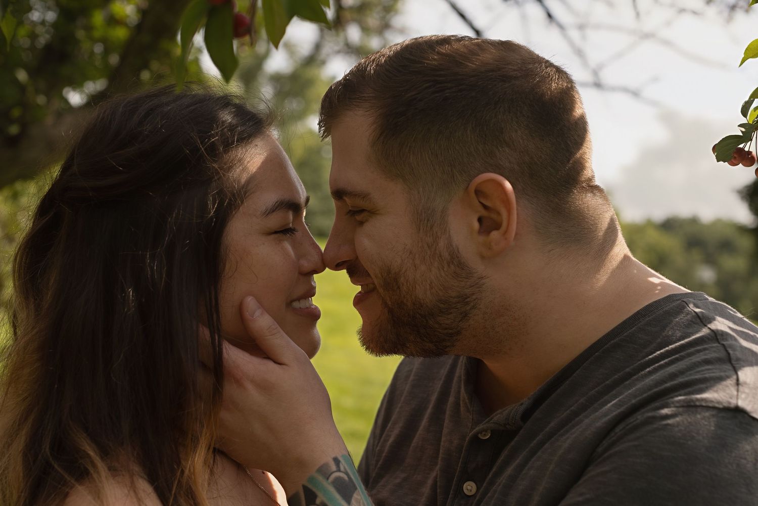 A romantic couple shares an intimate moment outdoors in warm sunlight against a natural background.