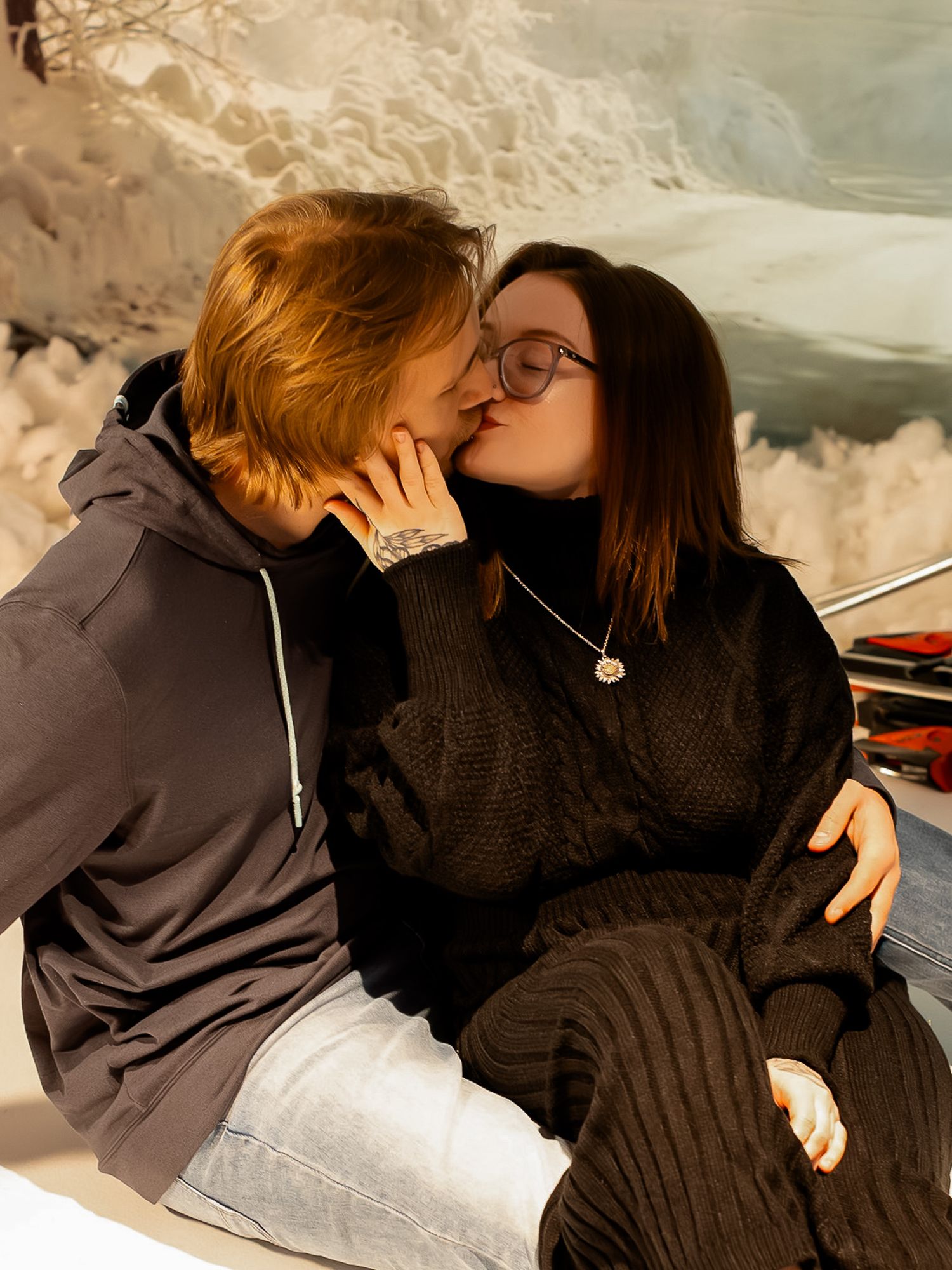 A romantic winter moment captured in warm sunlight as a couple shares an intimate kiss while sitting in the snow.