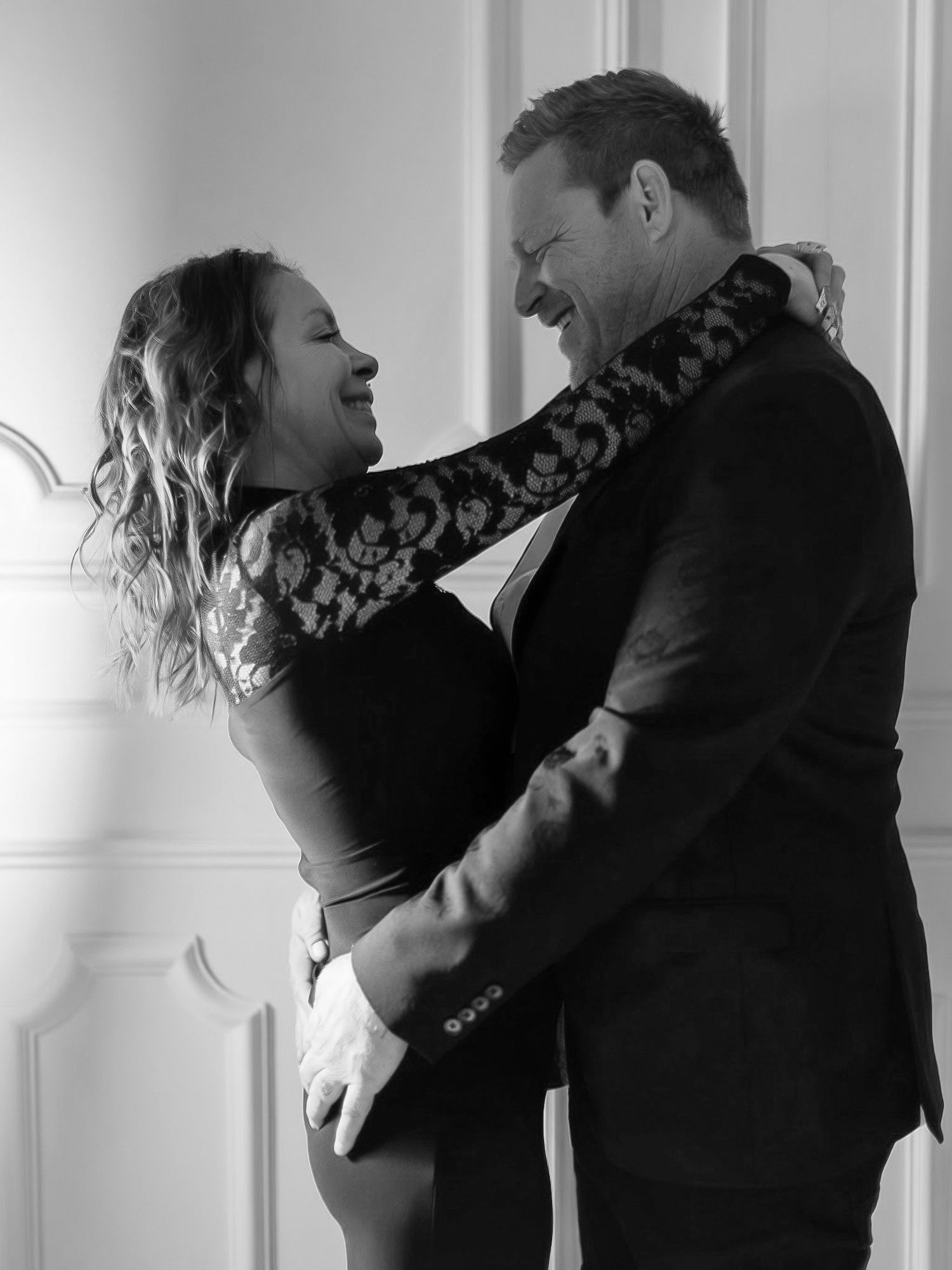 A romantic black and white portrait of a couple sharing an intimate embrace against a white wall.