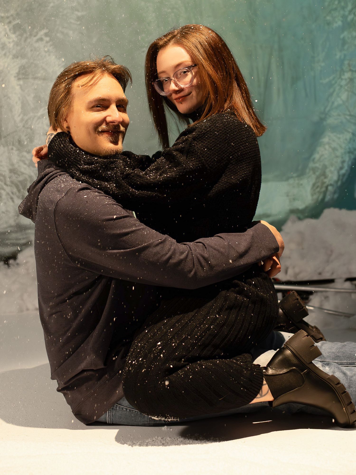 A playful winter portrait of a couple sitting together and smiling with snowflakes falling in the background.