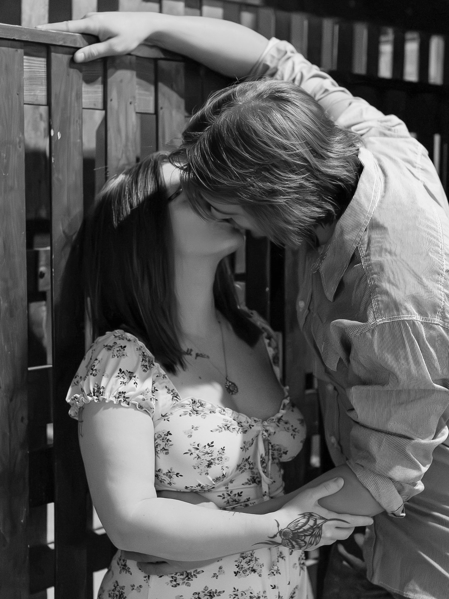 Black and white romantic photo of a couple in an intimate moment by a wooden fence, she wearing a floral dress.