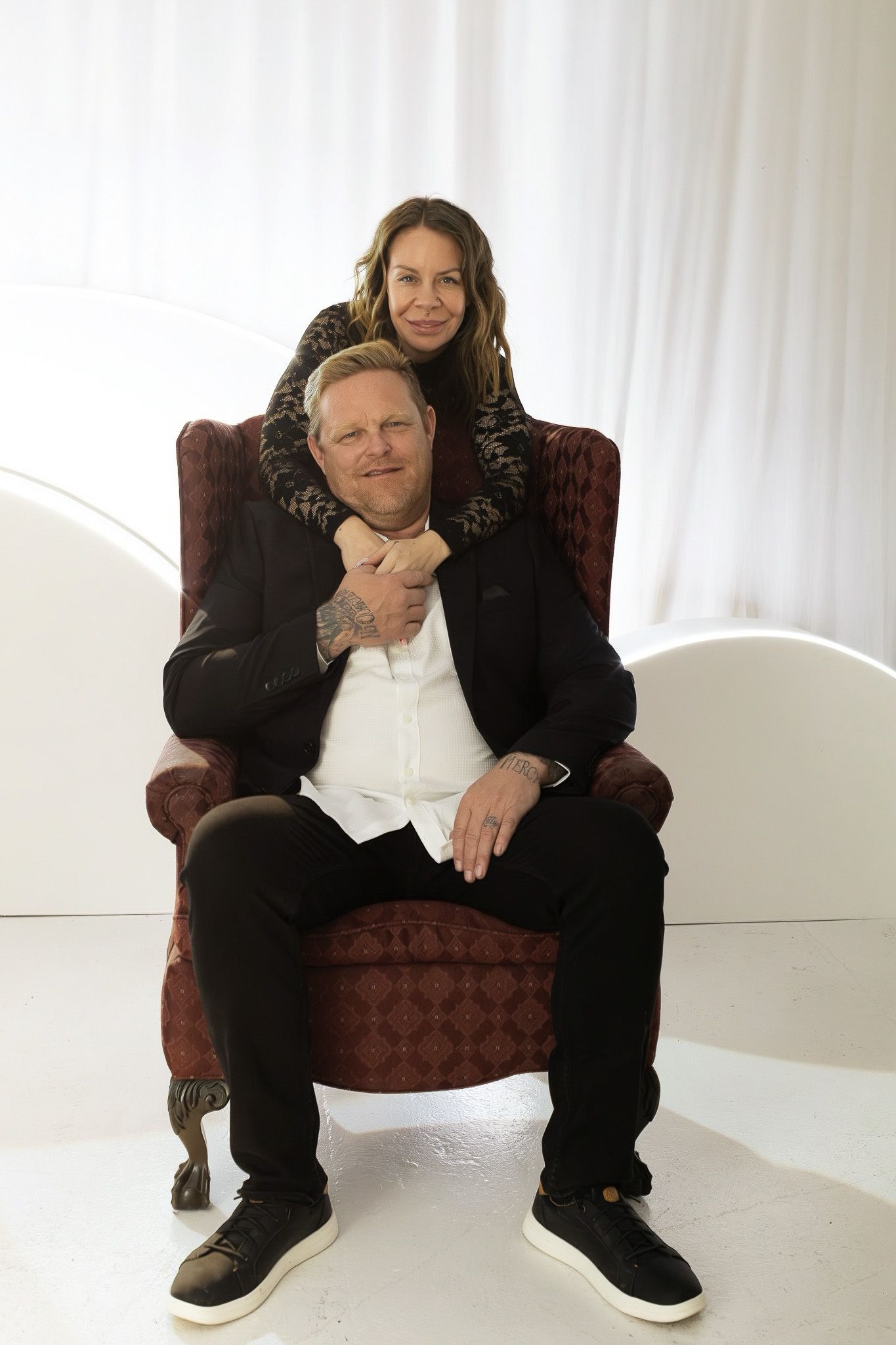 A couple poses together on a burgundy armchair in a bright white studio setting.
