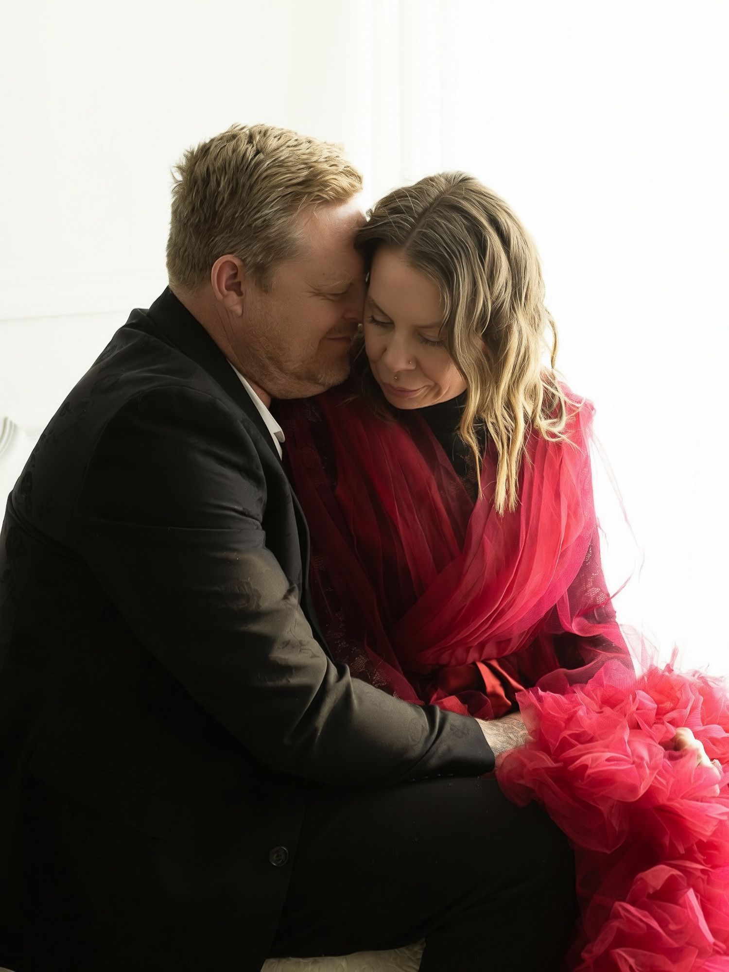Couple embraces tenderly with woman wearing dramatic red tulle gown during photoshoot.