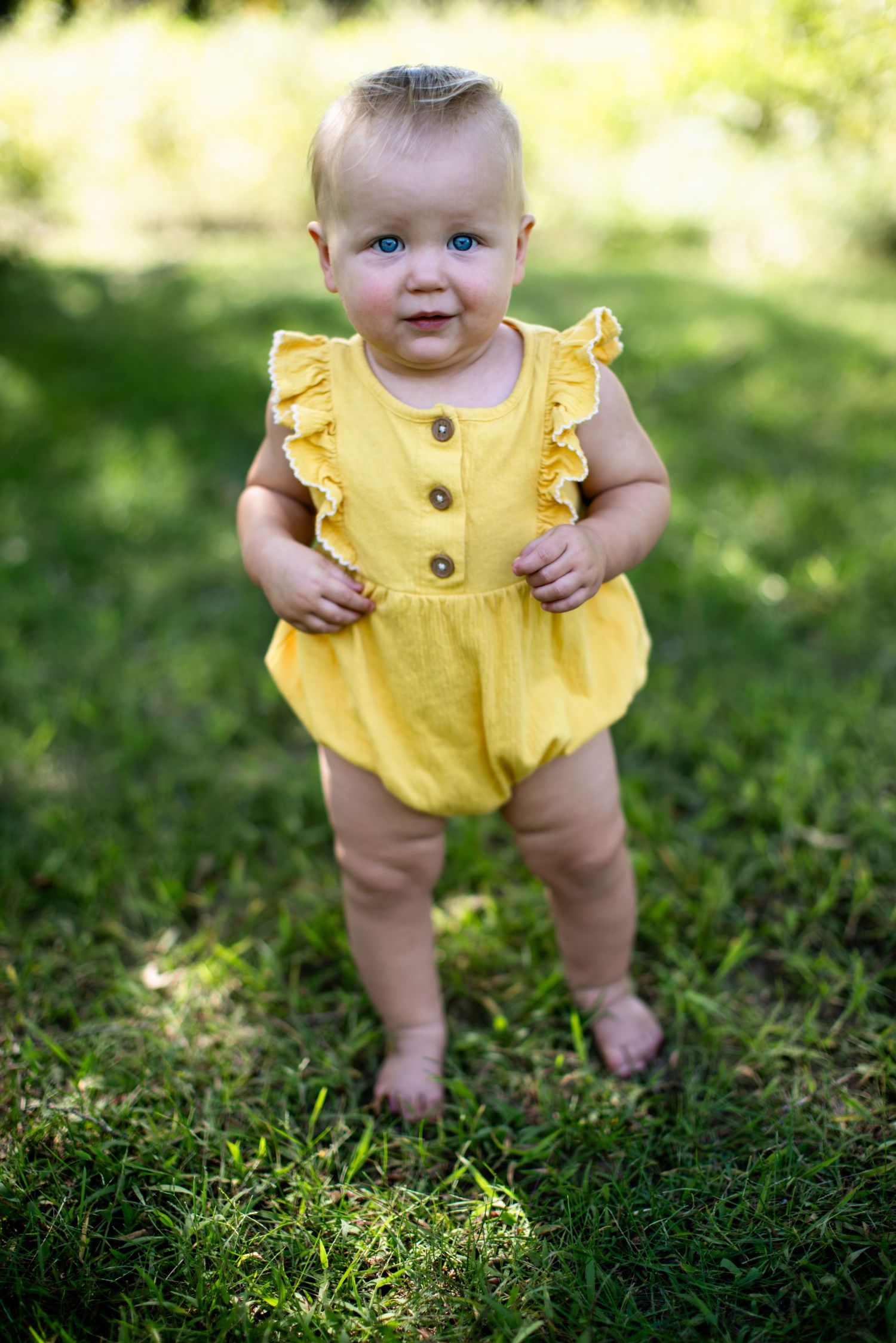 Baby in bright yellow ruffled romper standing on grass in sunlit garden.