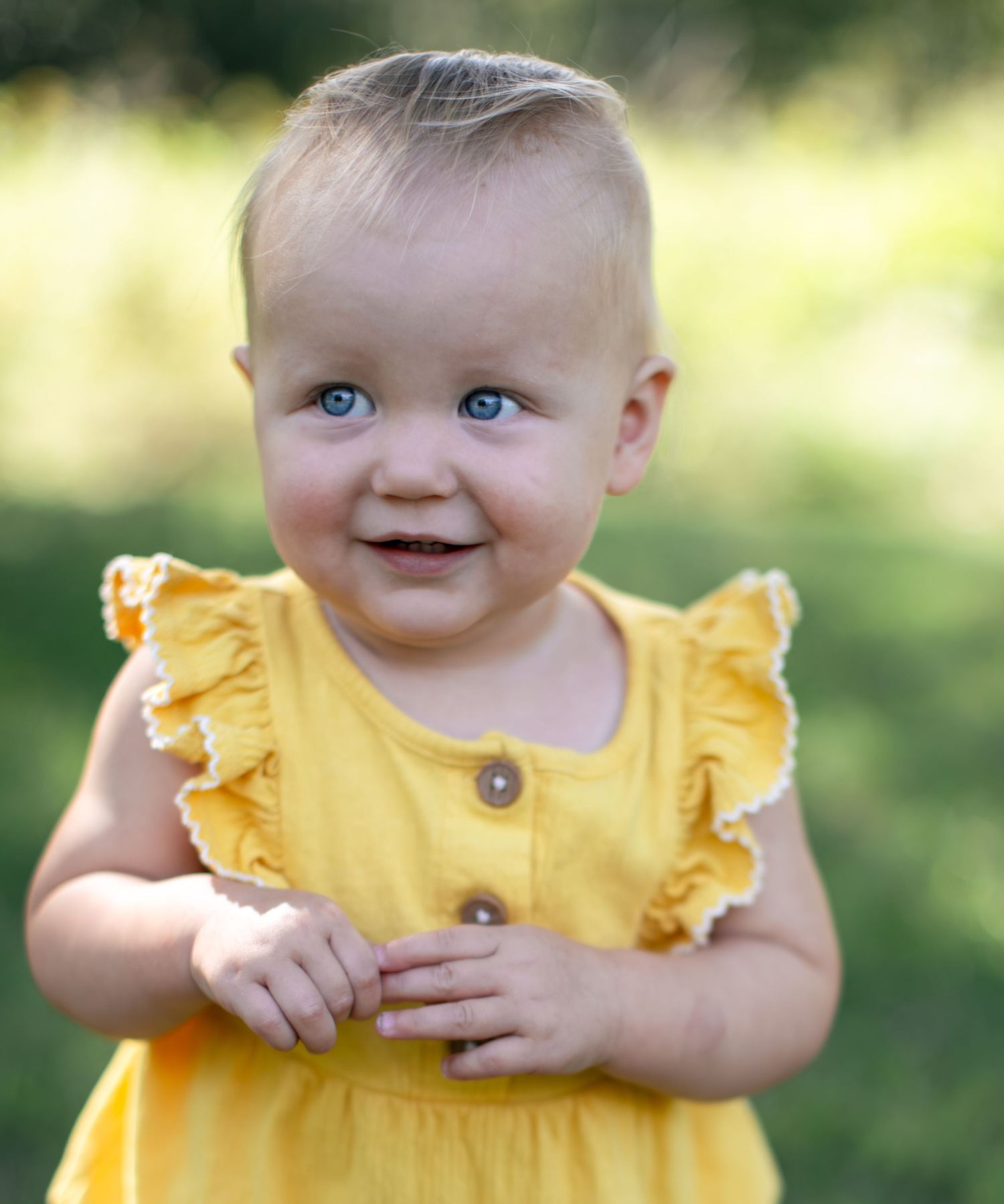 A baby wearing a bright yellow ruffled dress smiles while sitting outdoors in natural lighting.