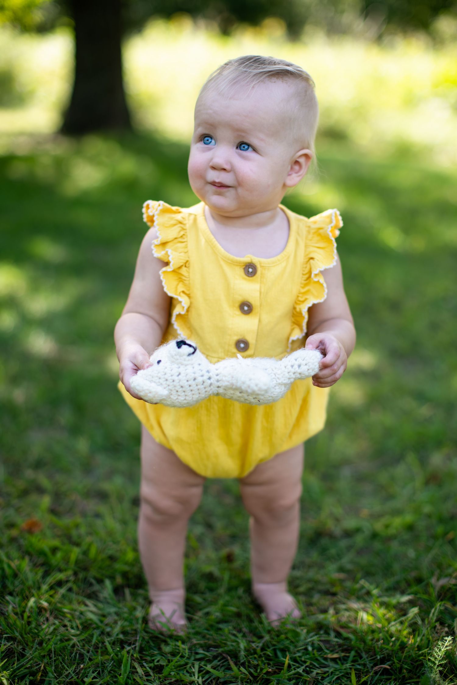 Small toddler wearing yellow ruffled romper holds white toy while standing on grass.