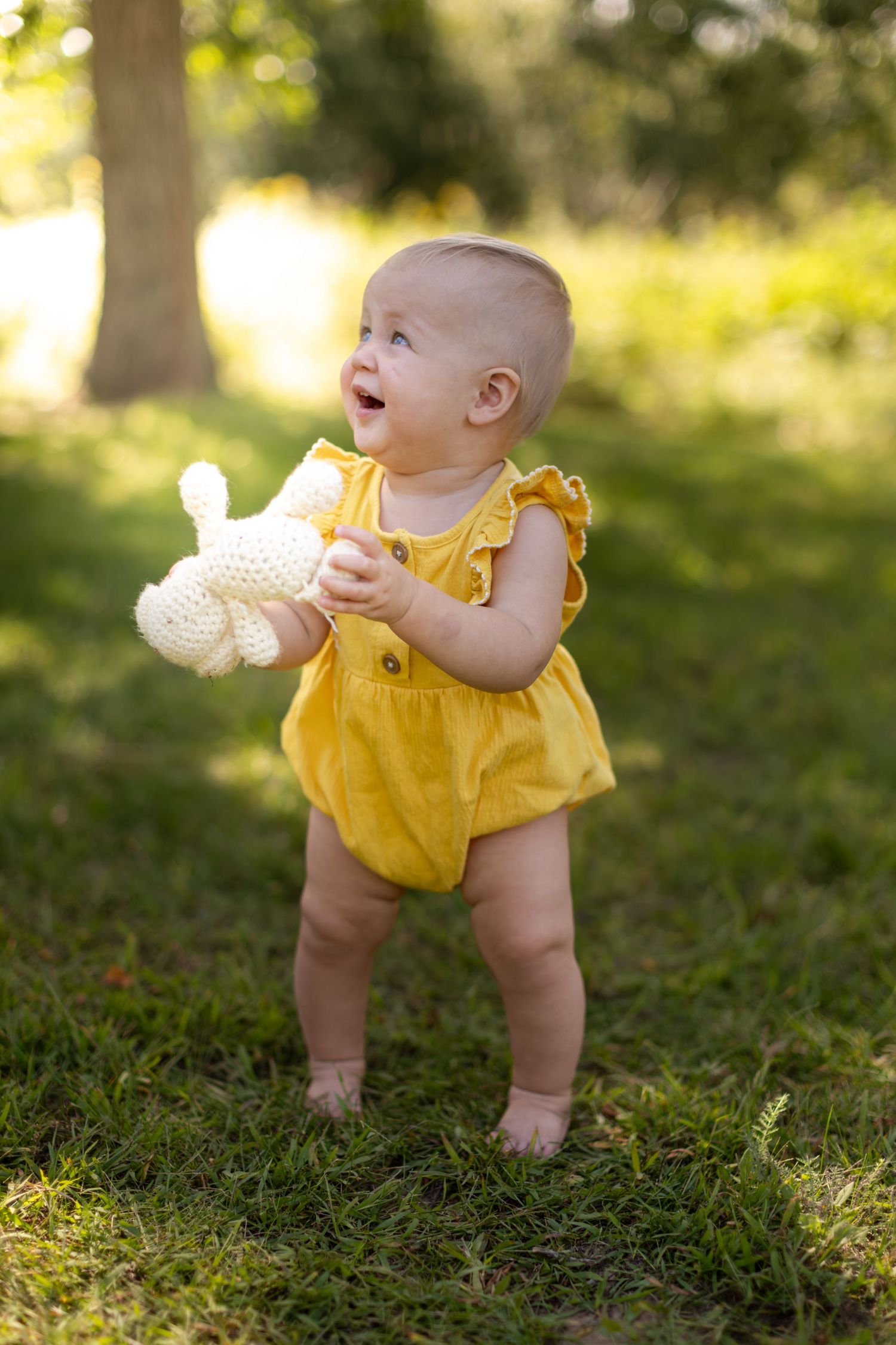 Baby in yellow outfit playing with white toy while sitting on grassy lawn.