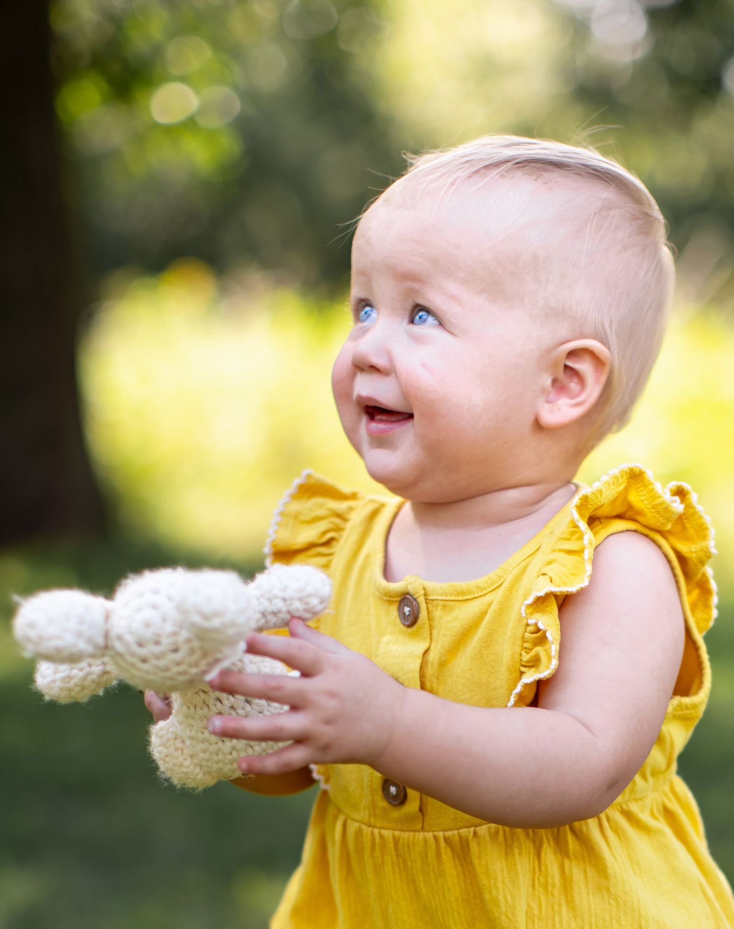 A small child in a yellow dress holds and plays with a white dandelion flower in a sunny outdoor setting.