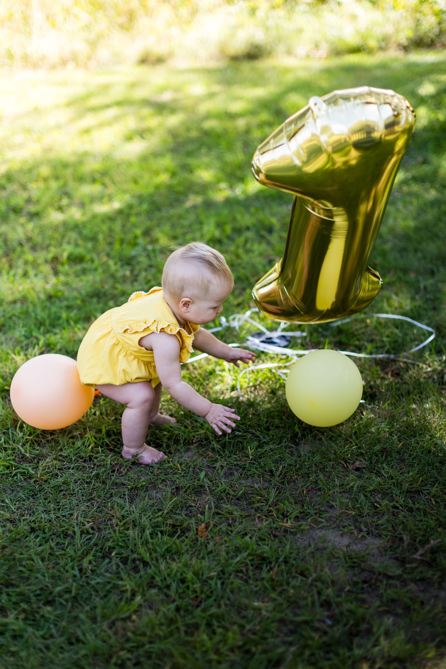 Baby in yellow dress playing with balloons on grass during golden hour.