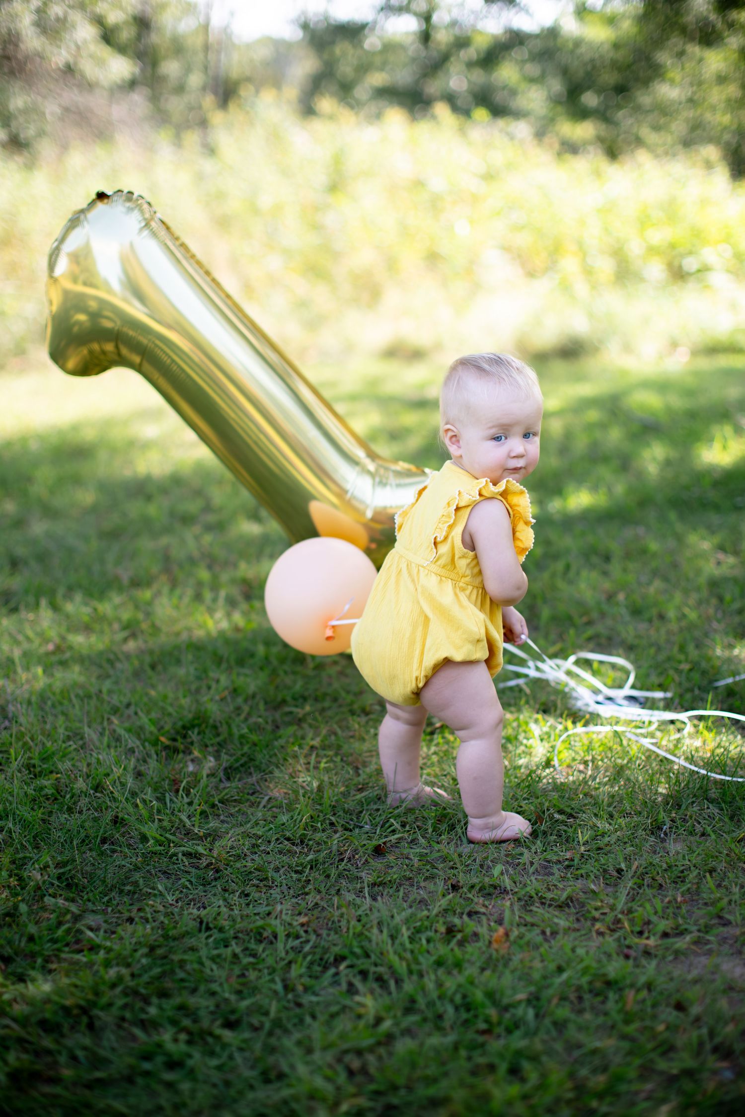 A baby in a yellow romper stands in a sunny field with balloons and a large gold number decoration.