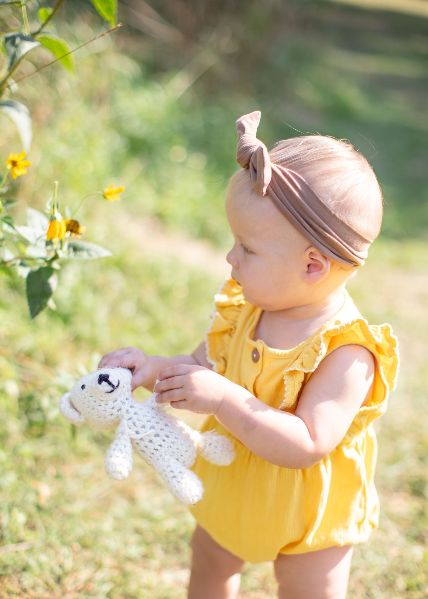 Small child in yellow outfit holding white plush toy in sunny outdoor setting.