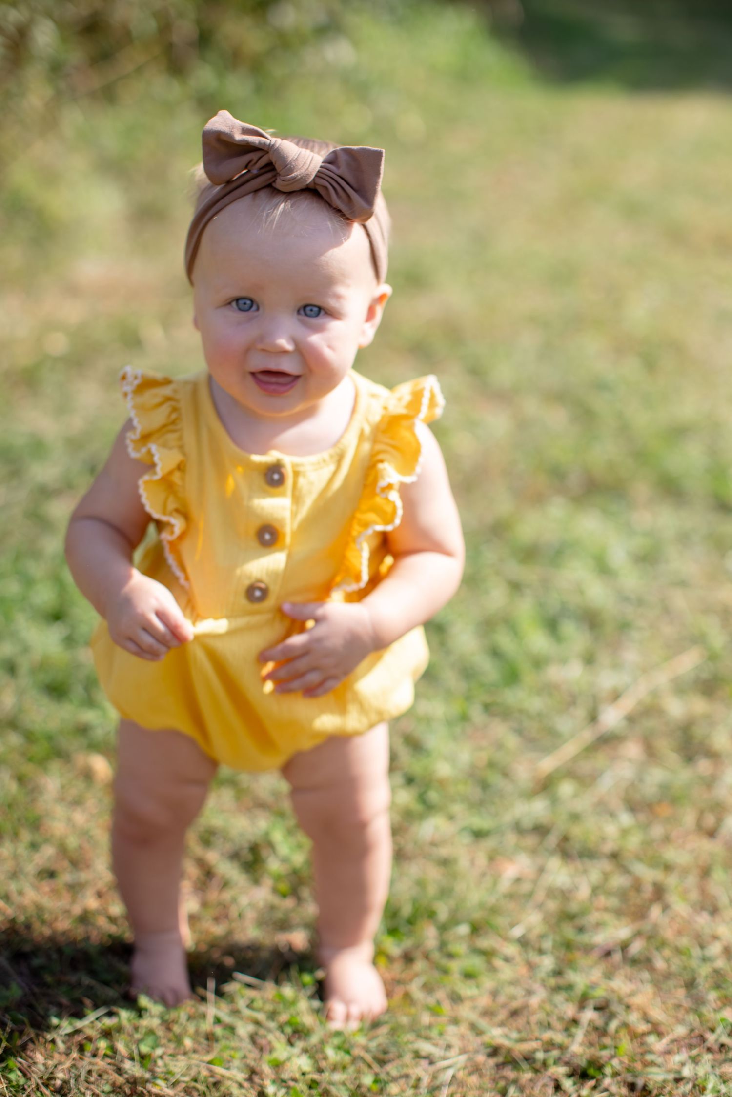 Happy baby in yellow ruffled romper and brown bow stands on grass in sunlit garden setting.