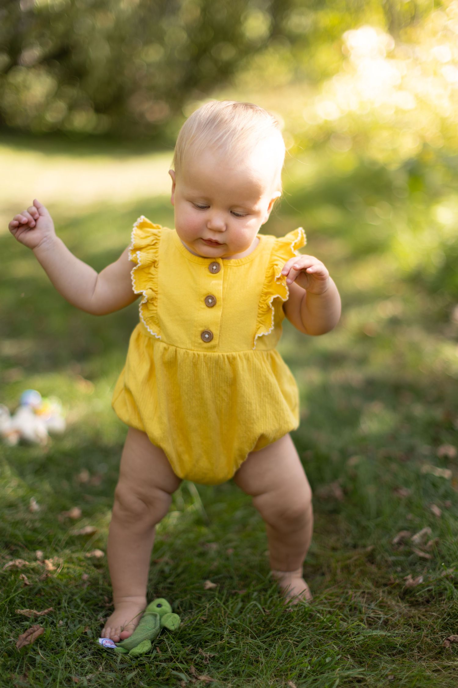 A baby wearing a ruffled yellow romper stands on grass in golden sunlight while dancing.