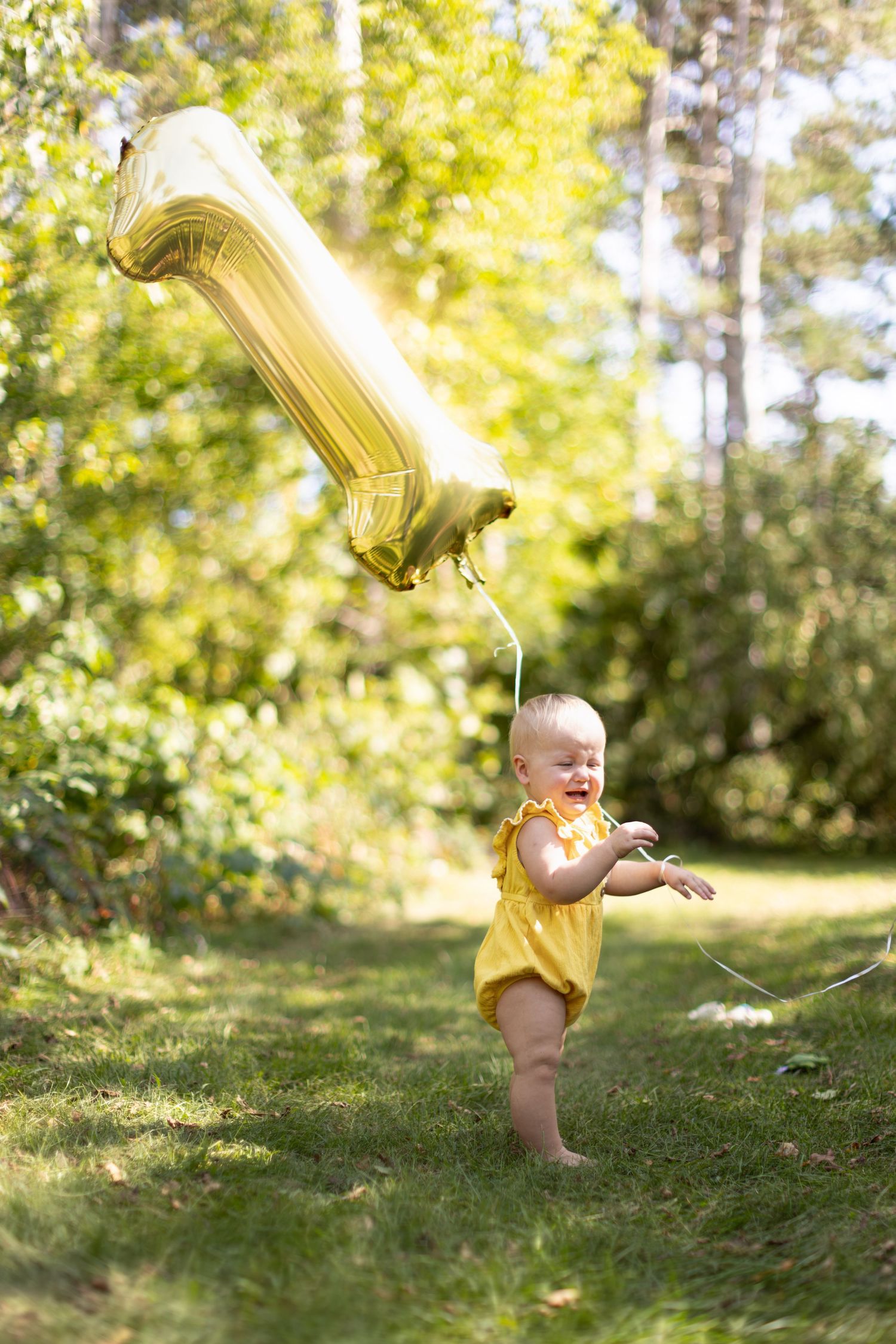 A baby in a yellow dress plays and crawls on green grass in a sunlit garden setting.