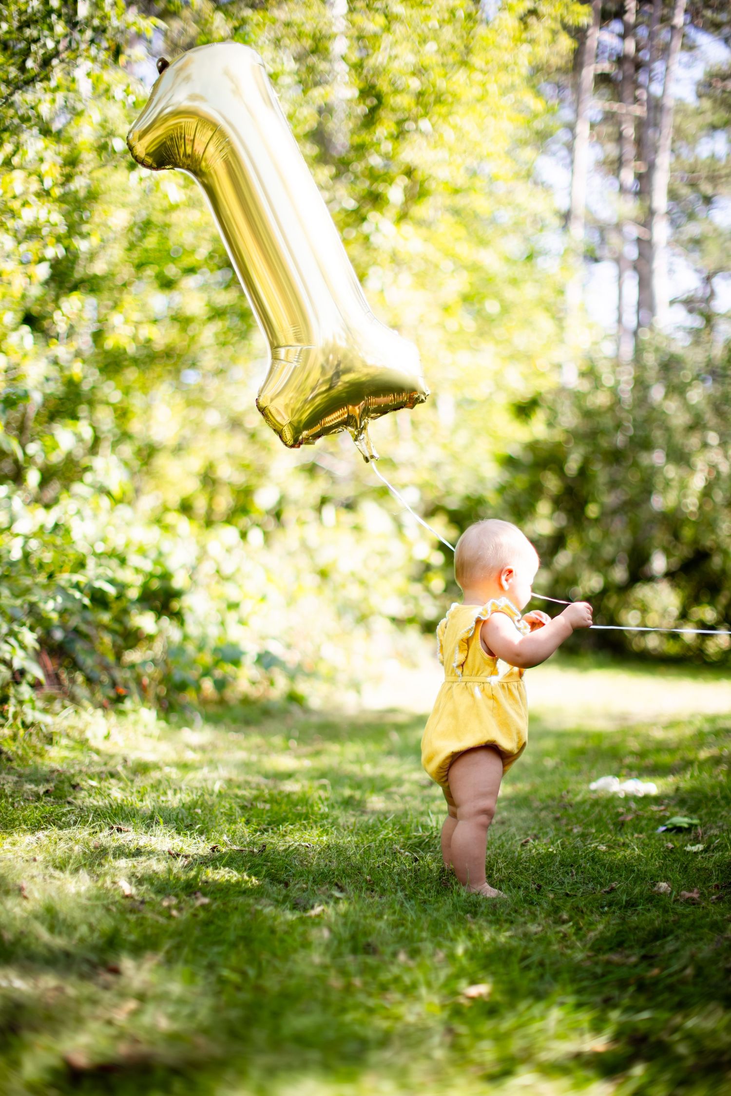 Baby in yellow outfit reaching up toward golden balloon in sunlit garden.