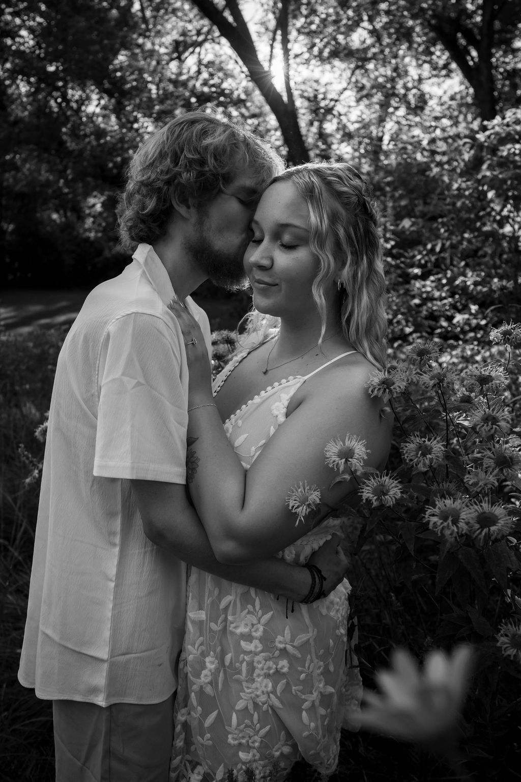 Black and white photo of a couple in a romantic embrace surrounded by flowering plants.