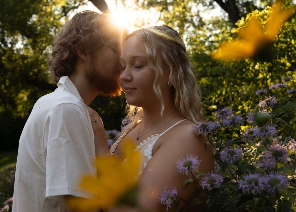 Sunlit garden scene with a couple sharing an intimate moment among yellow and purple flowers.