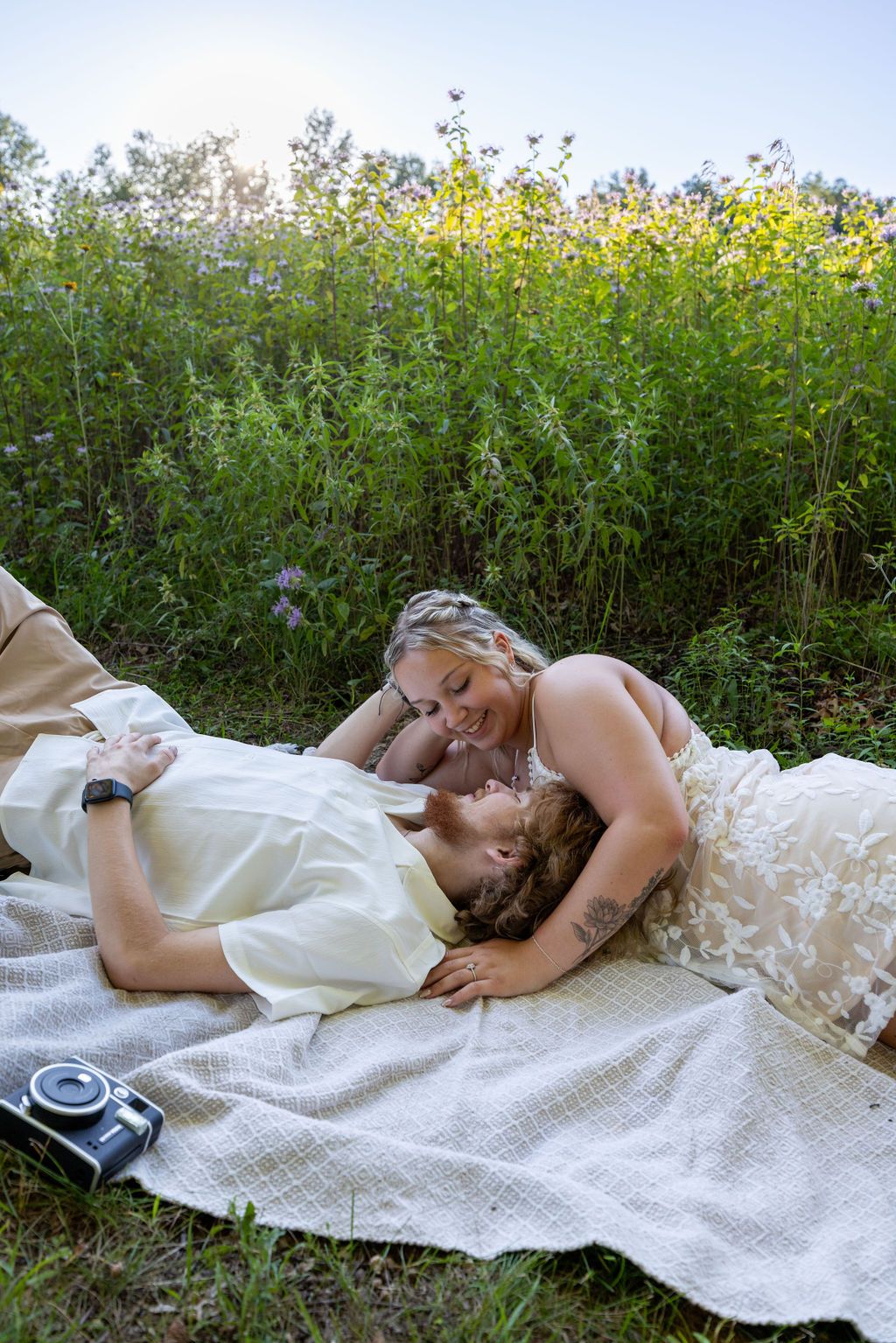 A couple lies together on a picnic blanket surrounded by tall wildflowers and greenery.