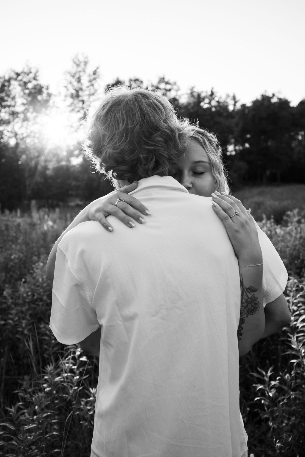 A tender embrace captured in black and white as sunlight filters through trees in a wildflower meadow.