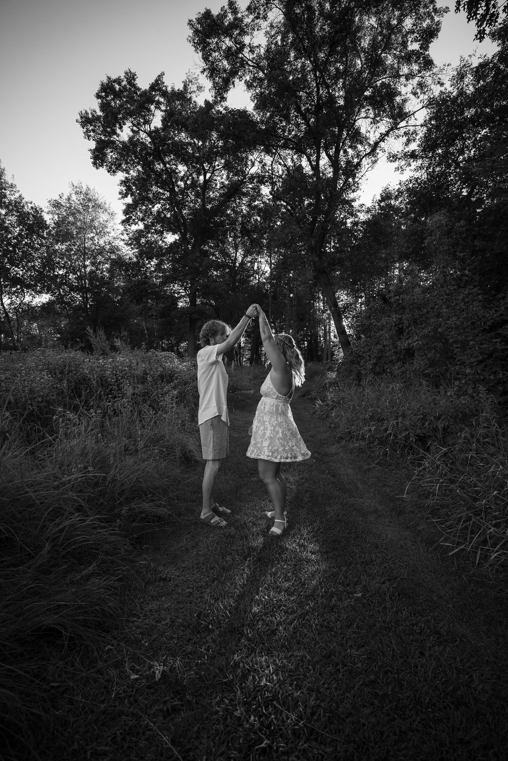 A monochrome shot of two people dancing together on a woodland path surrounded by tall grass.