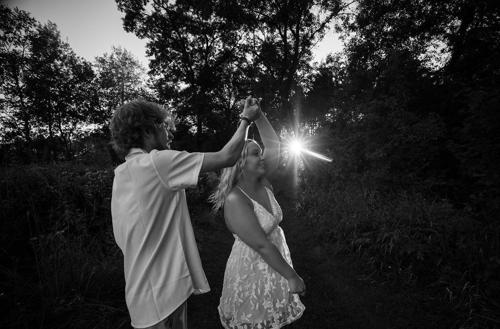 A black and white capture of two people dancing in nature with sunburst through trees.