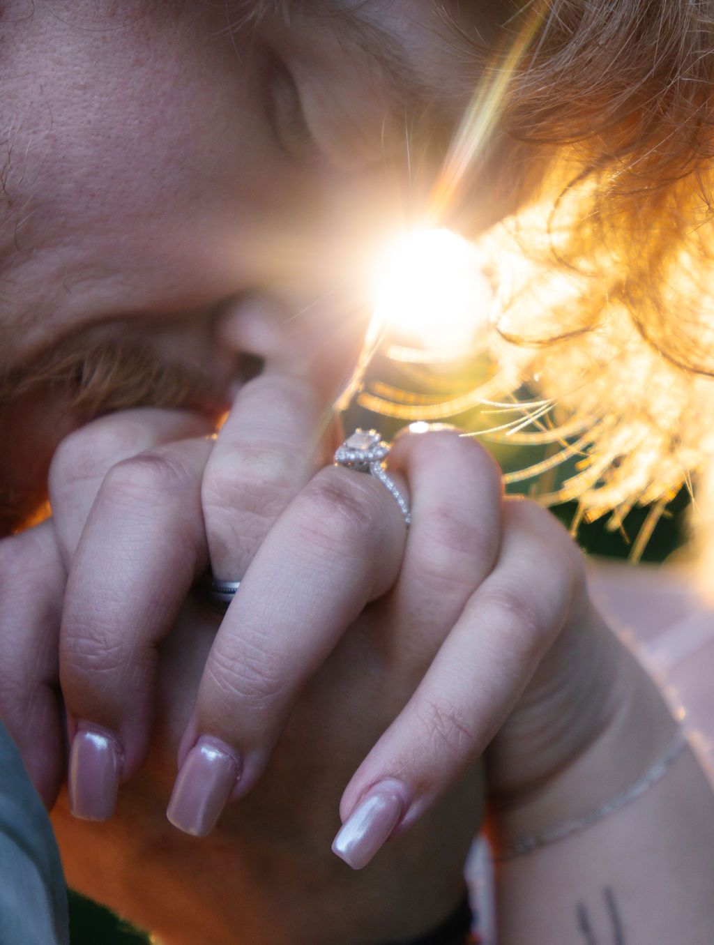 Sunlit close-up of manicured hands with engagement ring glowing in golden light.