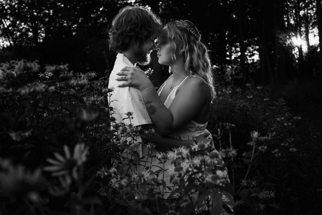 Intimate black and white portrait of couple embracing among wildflowers at sunset.