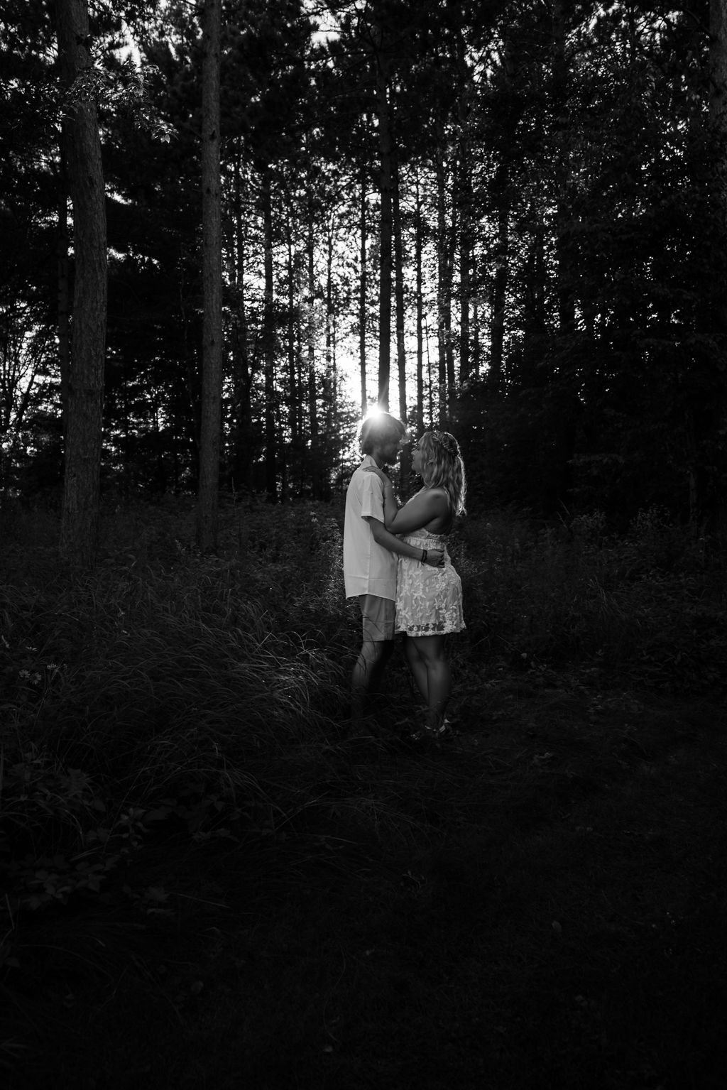 Black and white portrait of a couple sharing an intimate moment in a forest with sunlight streaming through trees.