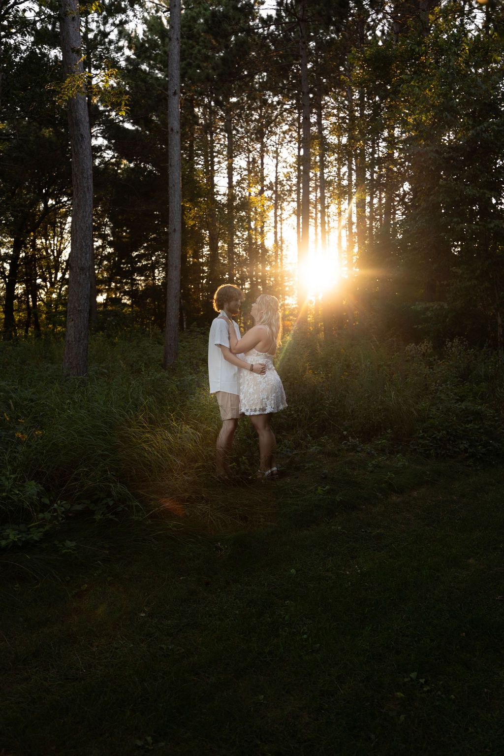 Couple in white clothing sharing a romantic moment in a sunlit forest during golden hour.