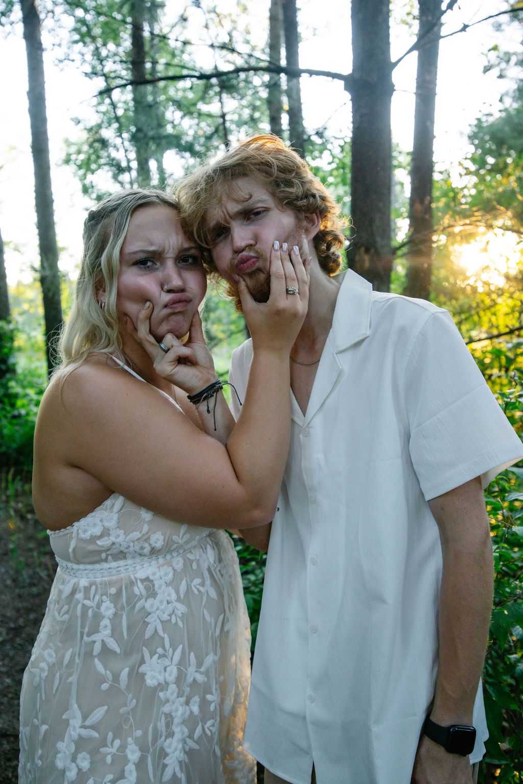 Playful couple making silly faces while posing for photos in a sunlit forest setting.