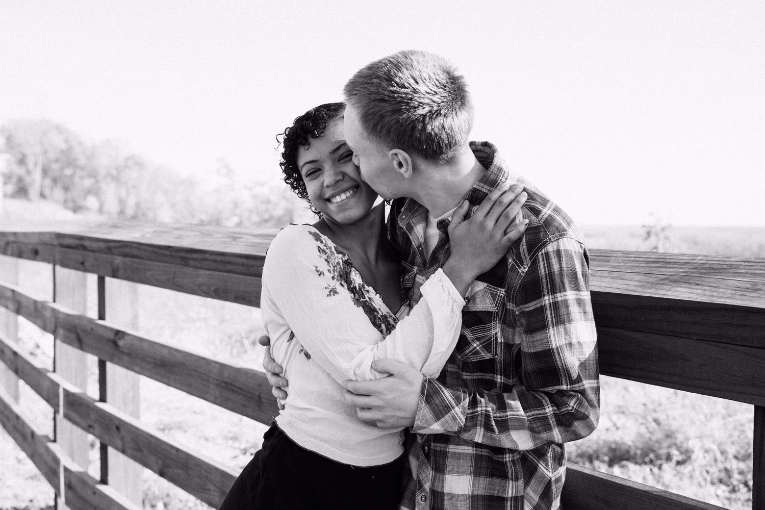 A black and white photo of a couple sharing a tender moment on a wooden deck overlook.