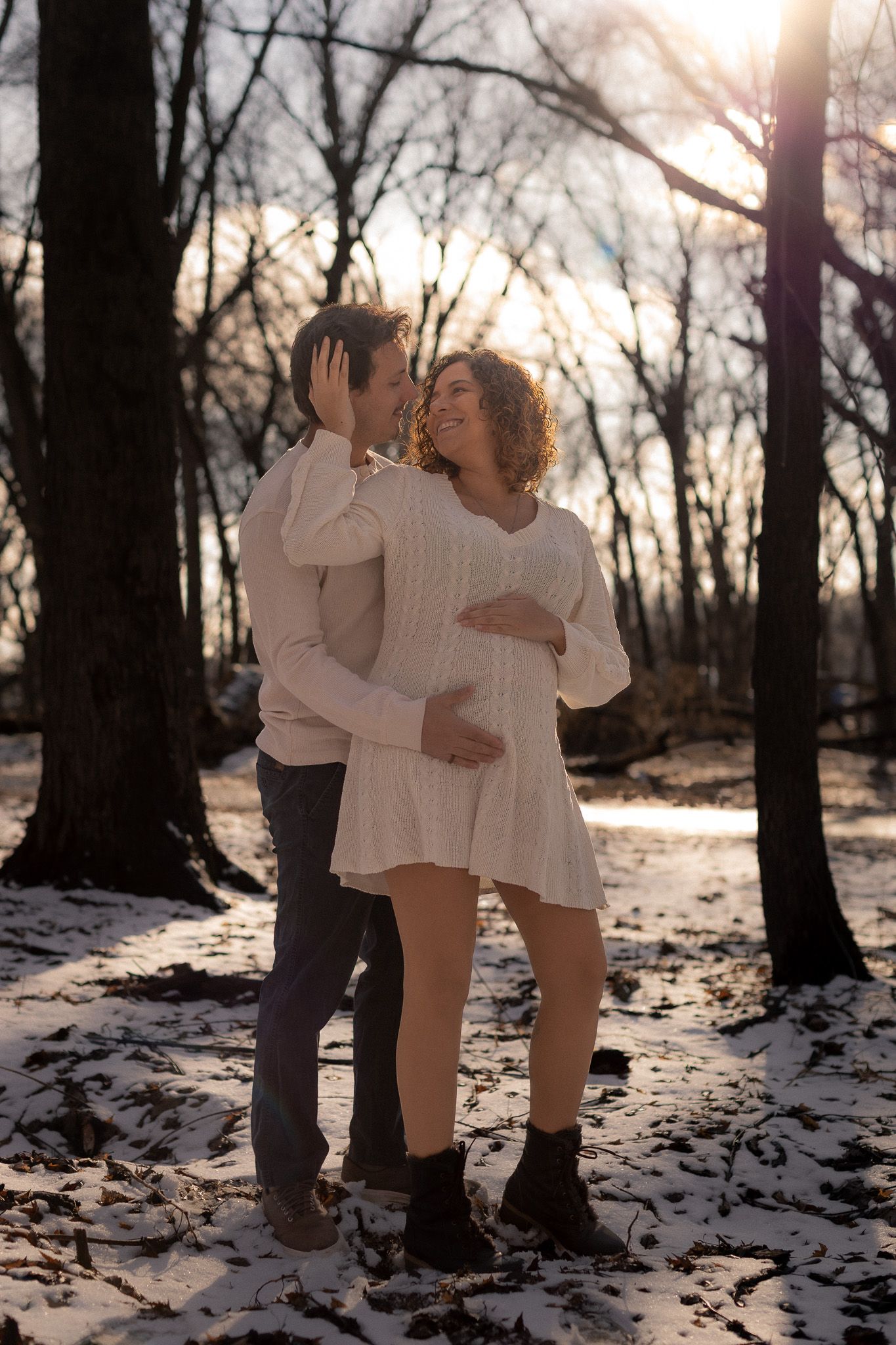 Two people in white outfits dance playfully together in a snowy forest at sunset.