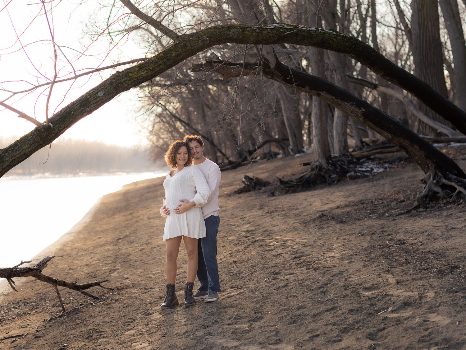 Couple in white outfits walking along a scenic beach at sunset with dramatic tree branches overhead.