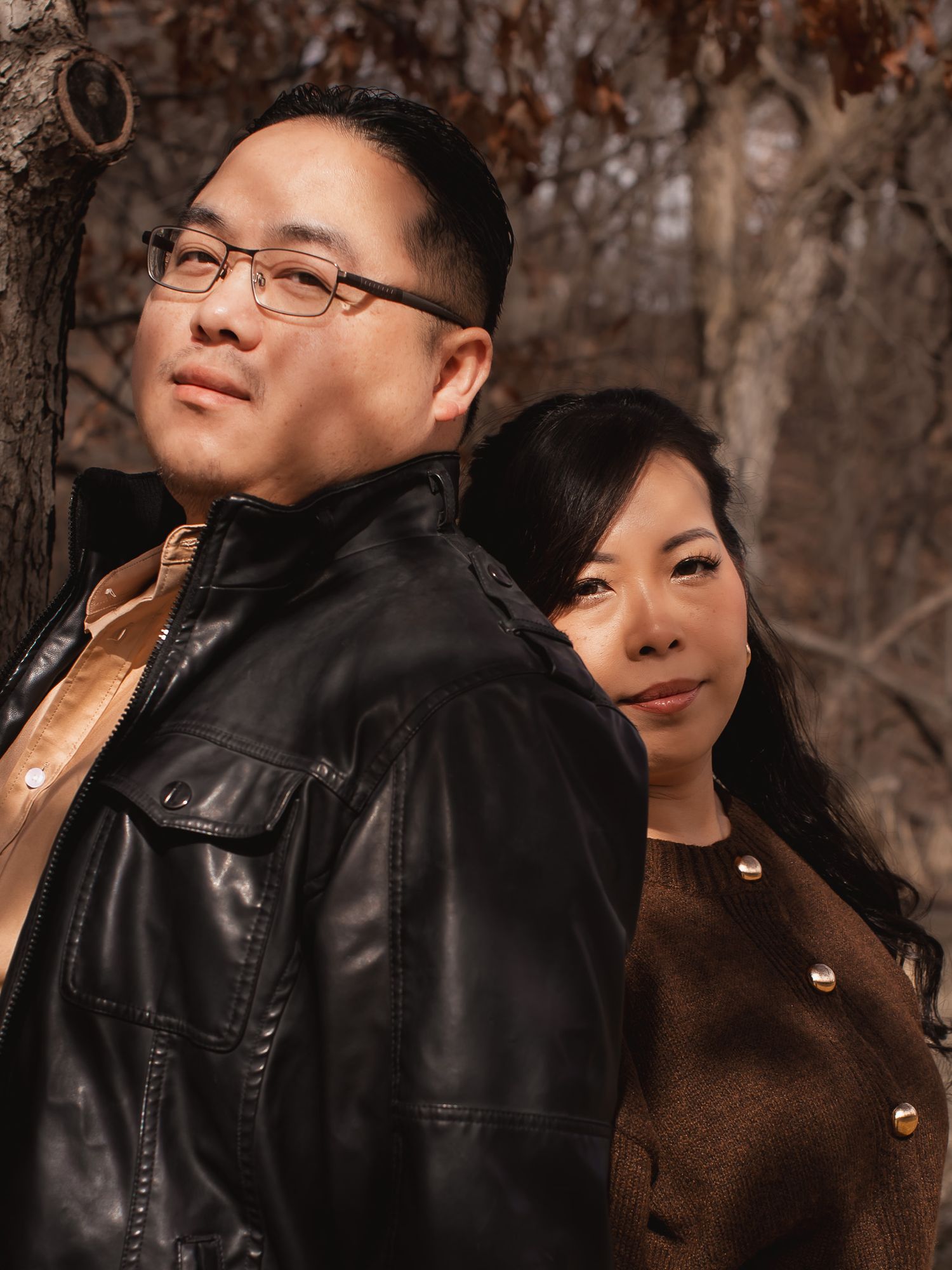 A couple in winter attire poses intimately together against a backdrop of bare trees in a romantic outdoor setting.