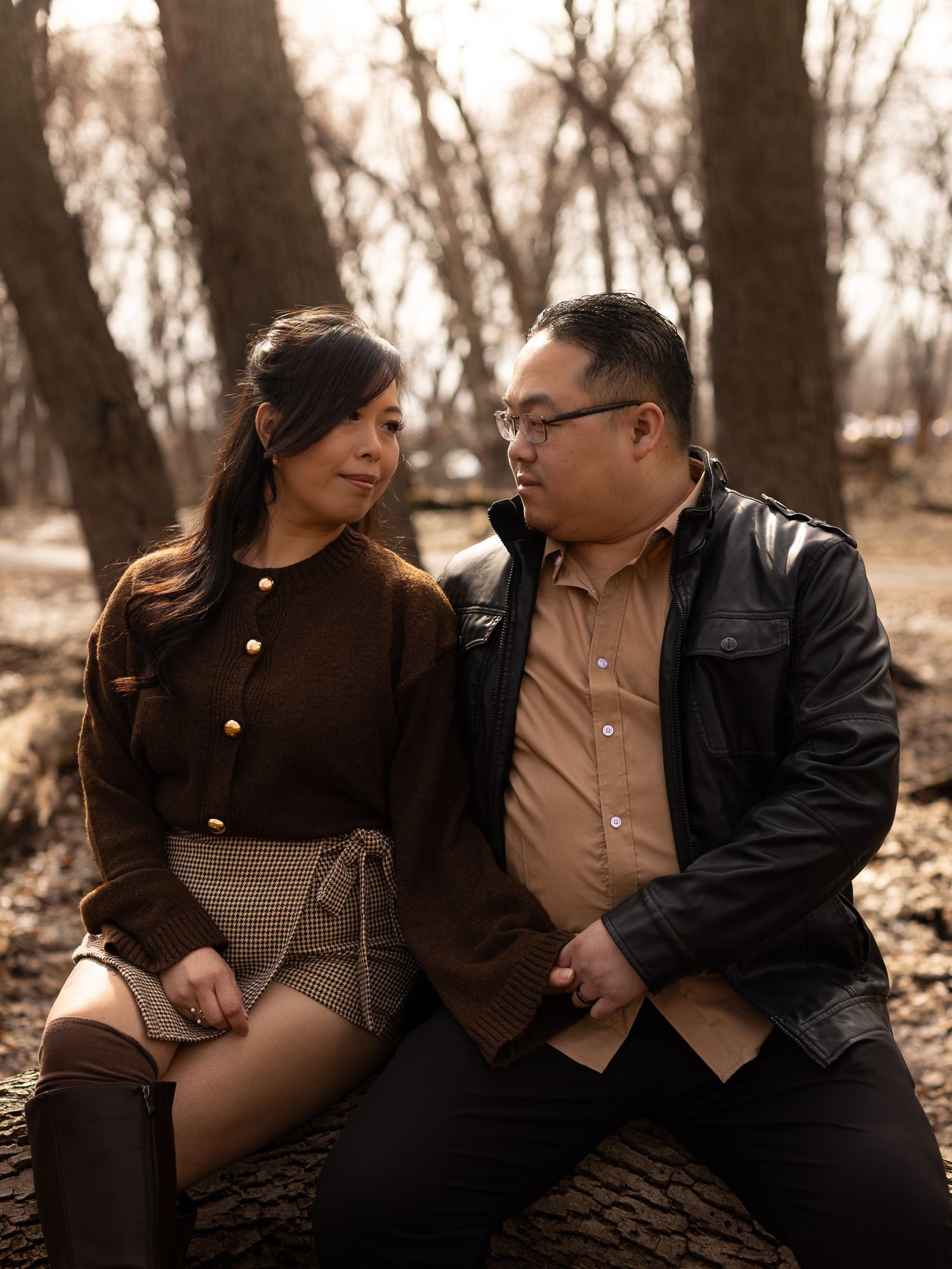 A romantic couple sits close together on a fallen tree trunk in a wooded area during autumn.