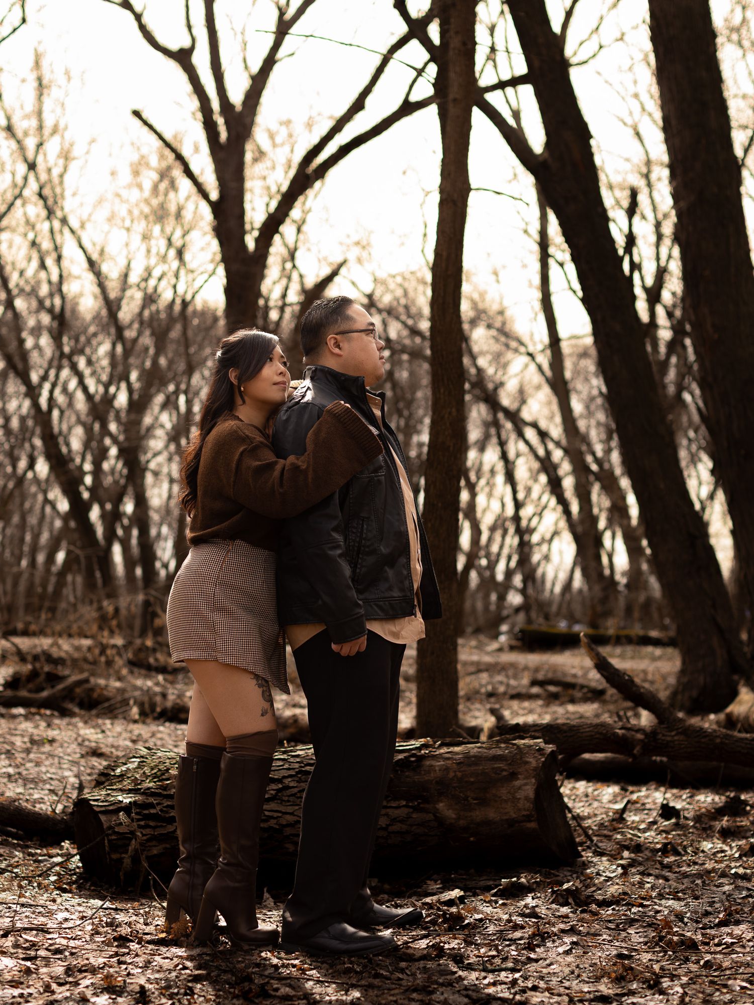 Romantic couple embraces in a wooded area during sunset with trees silhouetted in the background.