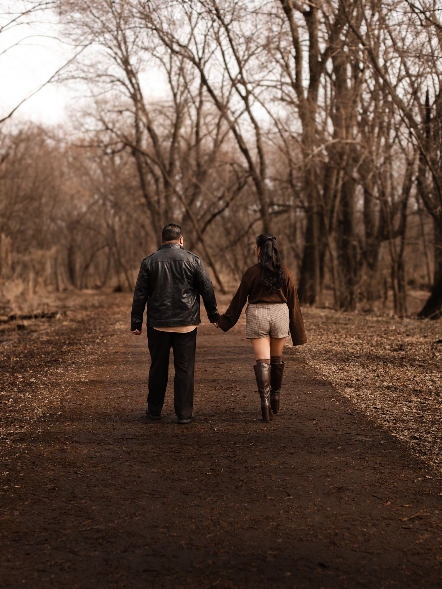 A couple holding hands while walking on an autumn path through bare trees in sepia tones.
